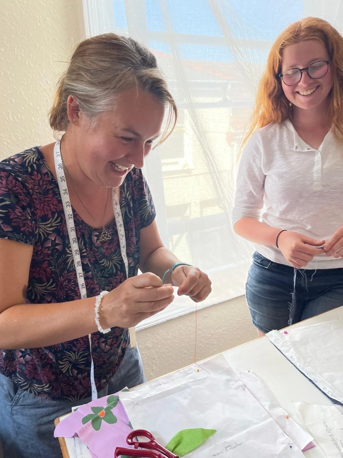 Two women are working together at a table with sewing materials, smiling and laughing. One woman has gray hair and glasses, and the other has red hair and glasses.