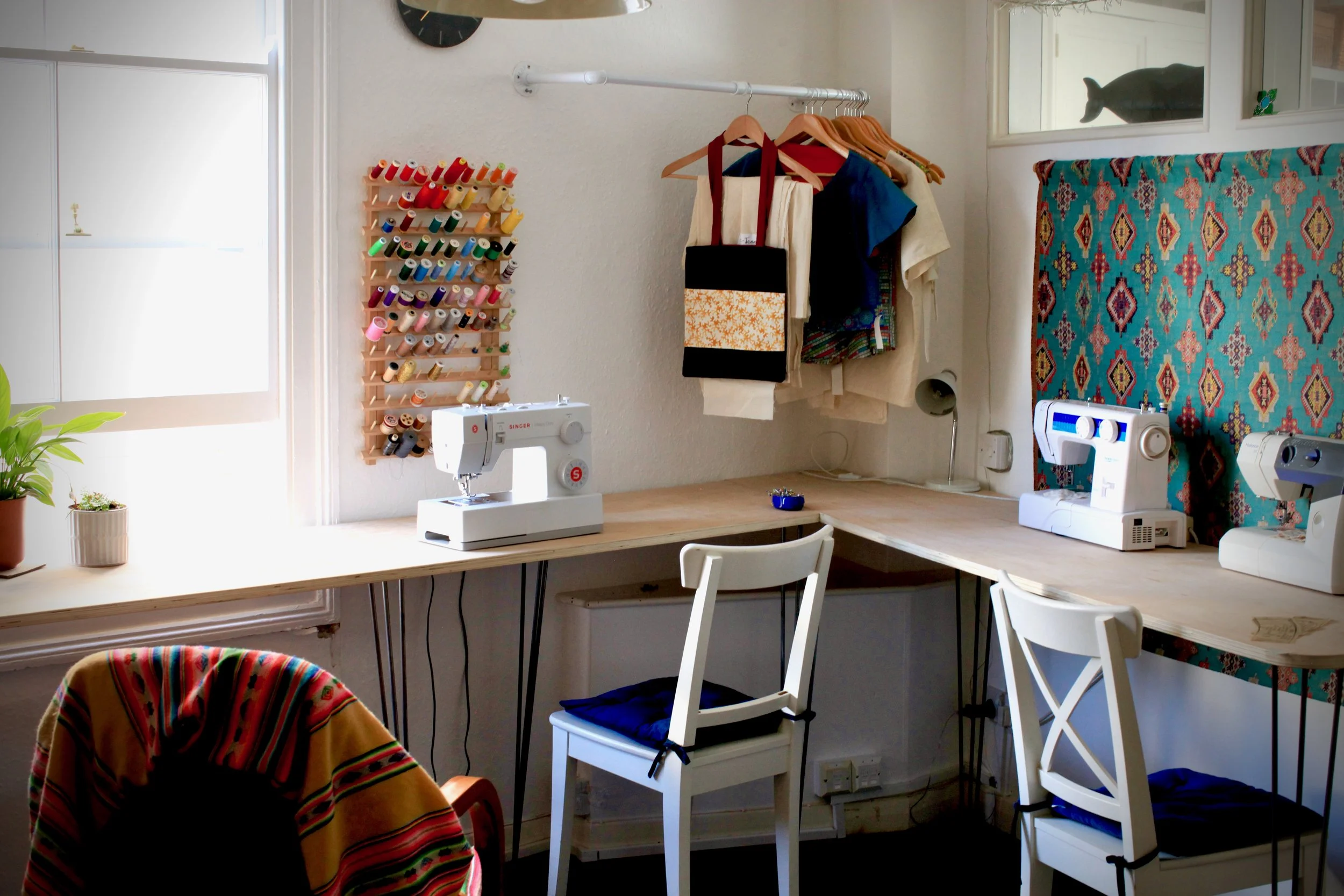 A sewing room with a wooden work table, sewing machines, white chairs, a window, colorful fabric, and yarn spools on a wall display.