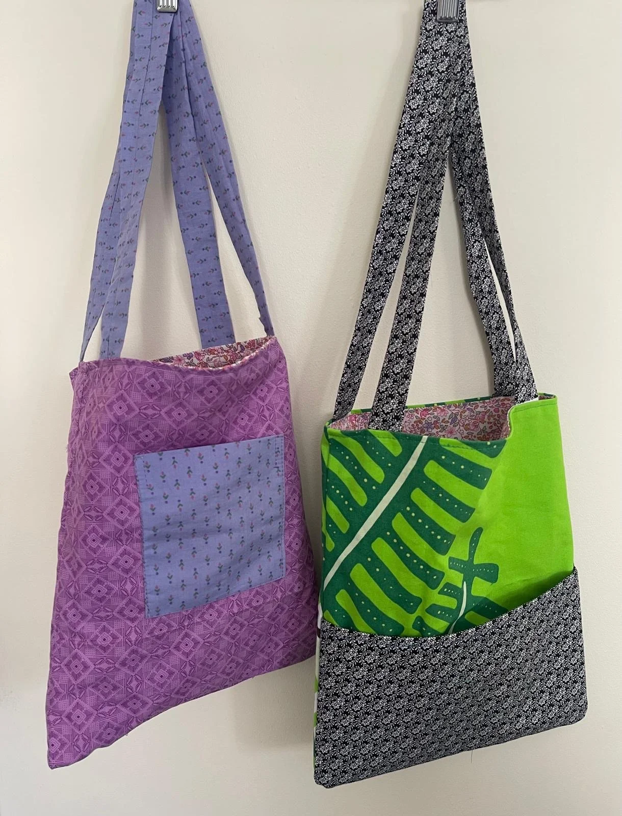 Two fabric tote bags hanging on a wall, one purple with a square patch and lavender patterned straps, the other green with a leaf design and black and white patterned bottom, with black and white patterned straps.