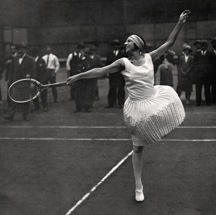 A woman in vintage tennis attire, including a pleated skirt and headband, playing tennis on a court with spectators in the background.