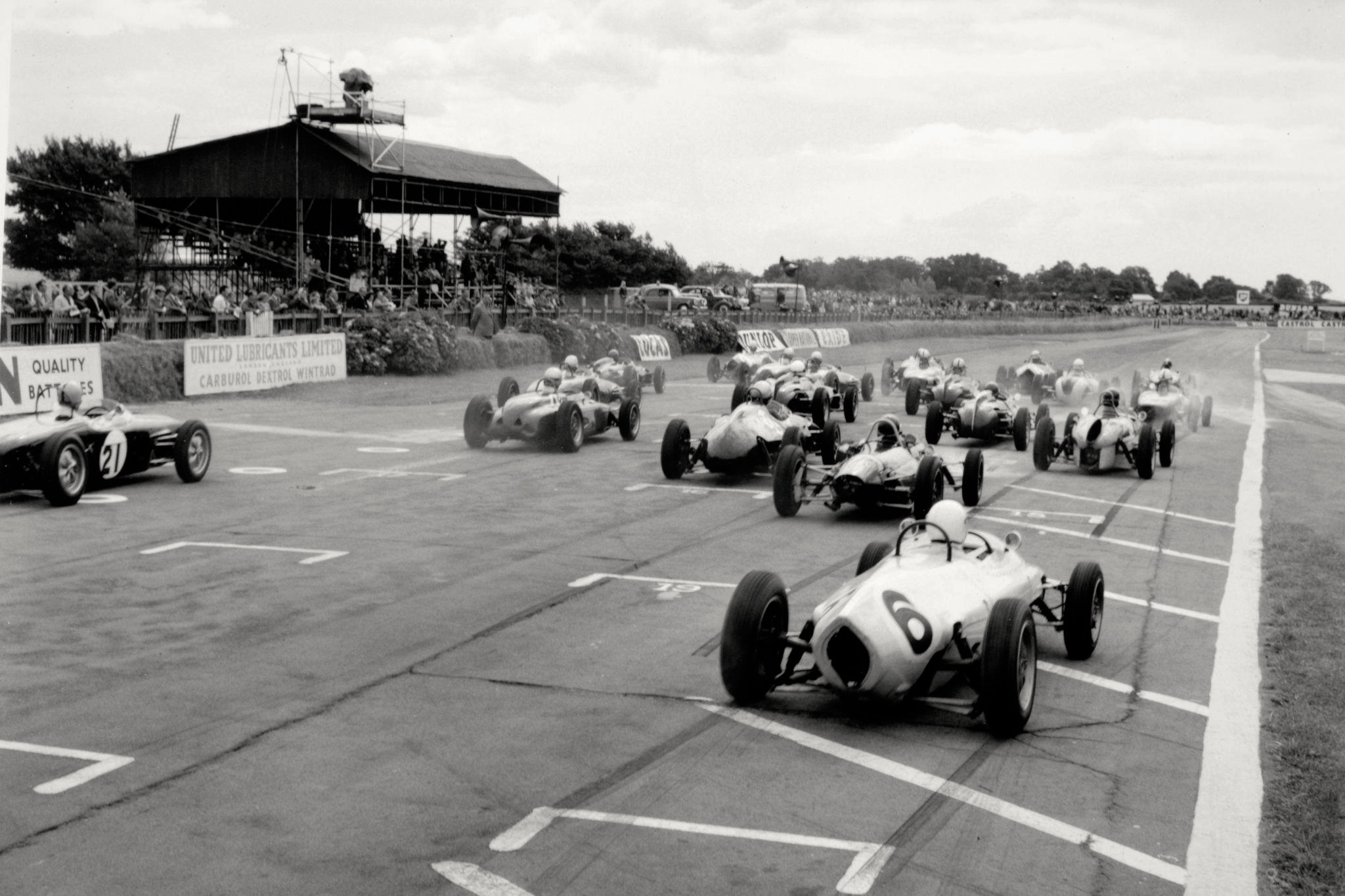 Vintage black-and-white photo of a racing event with multiple open-wheel race cars moving on a track, spectators seated in stands, and a banner advertising lubricants.