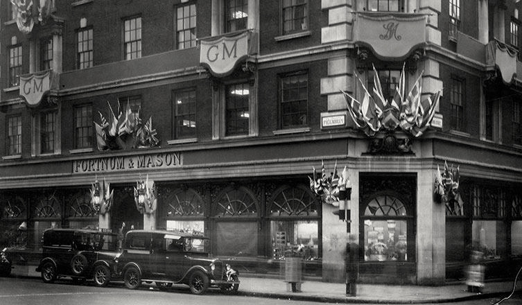 Black and white photo of the Fortnum & Mason store in London with flags and signs, parked vintage cars, and people walking on the street.