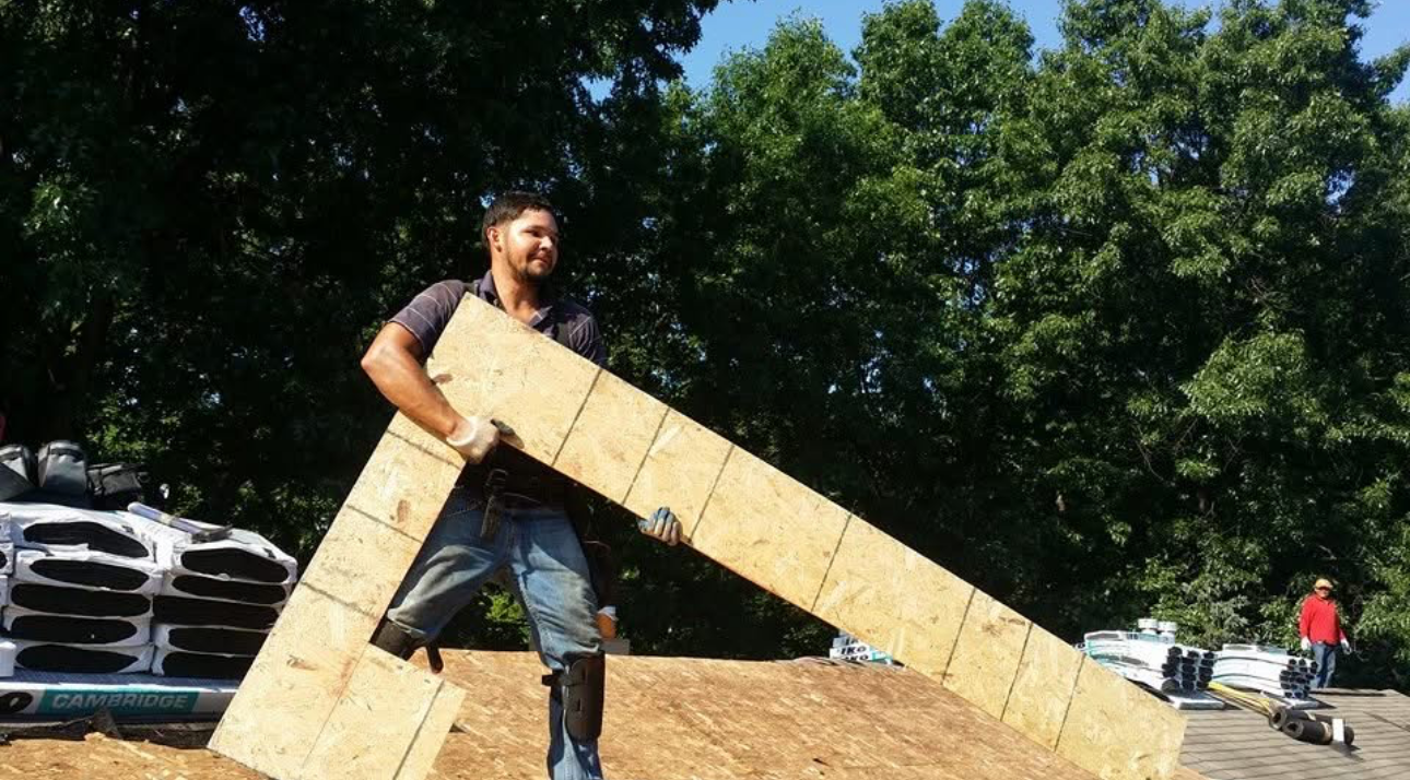 Man holding a large piece of construction material outdoors on a construction site with trees in the background.