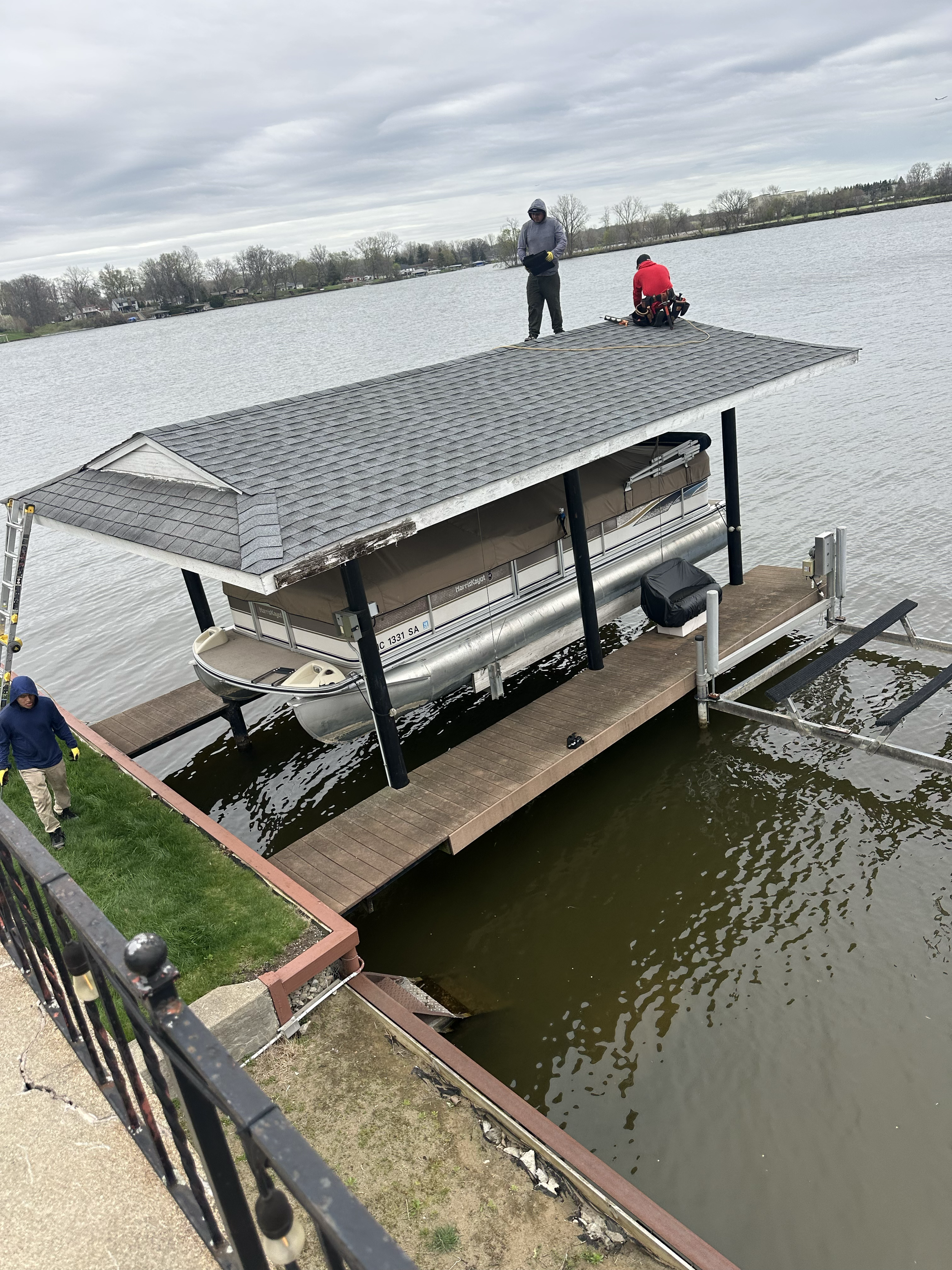 Two workers on a dock repairing or inspecting a covered pontoon boat in a body of water during overcast weather. One worker is standing, and the other is kneeling. The boat is moored to the dock with a ladder and railing nearby.