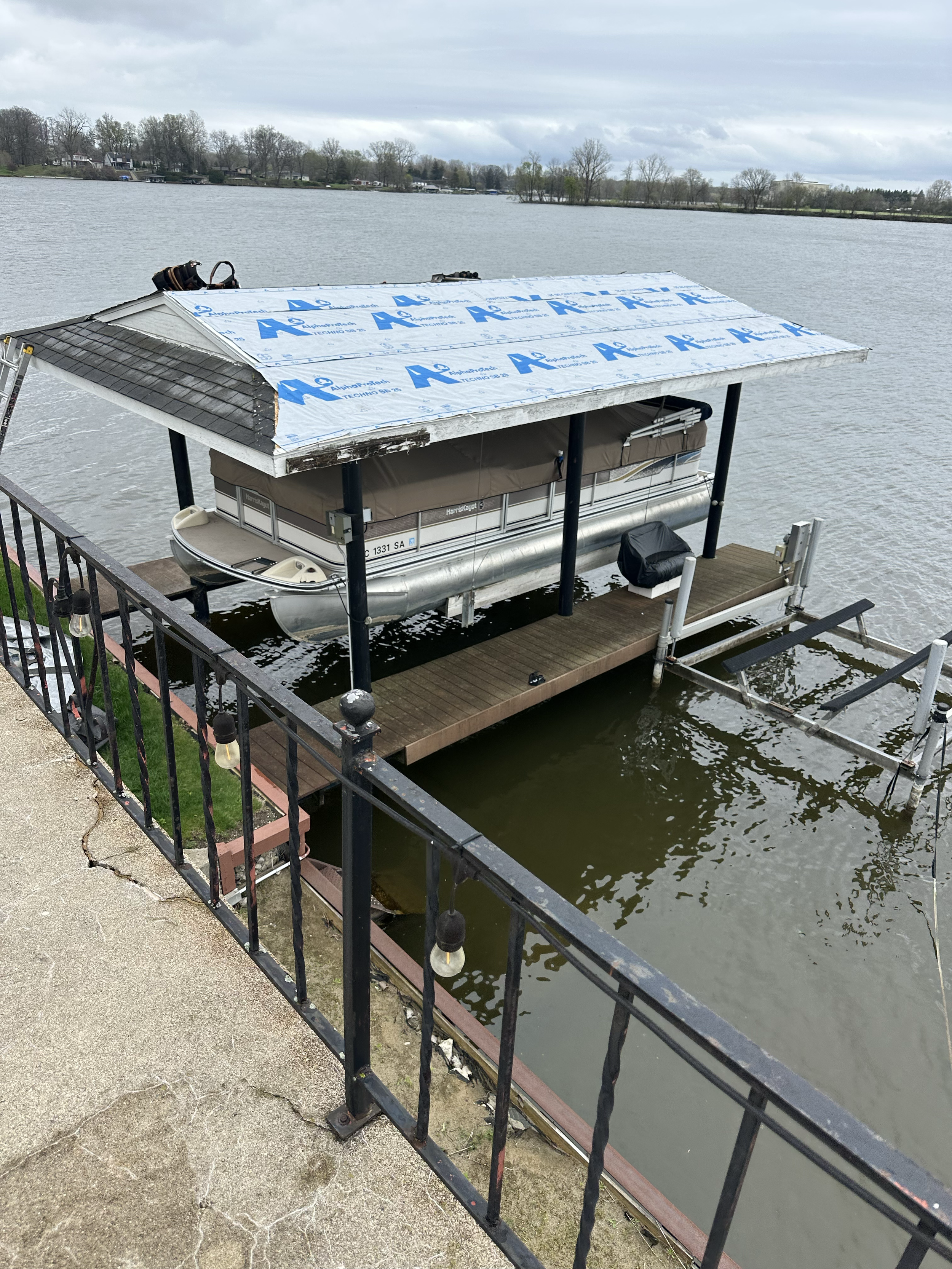 A pontoon boat docked under construction on a lake, with a partially installed roof and a railing around the dock, overcast sky in the background.