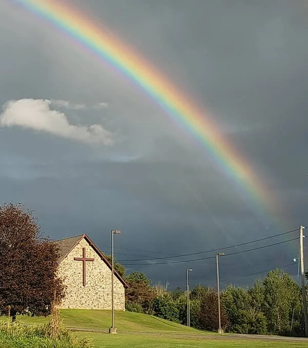 A rainbow arching in a cloudy sky over a church with a cross on the front, surrounded by trees and sidewalk street lights.