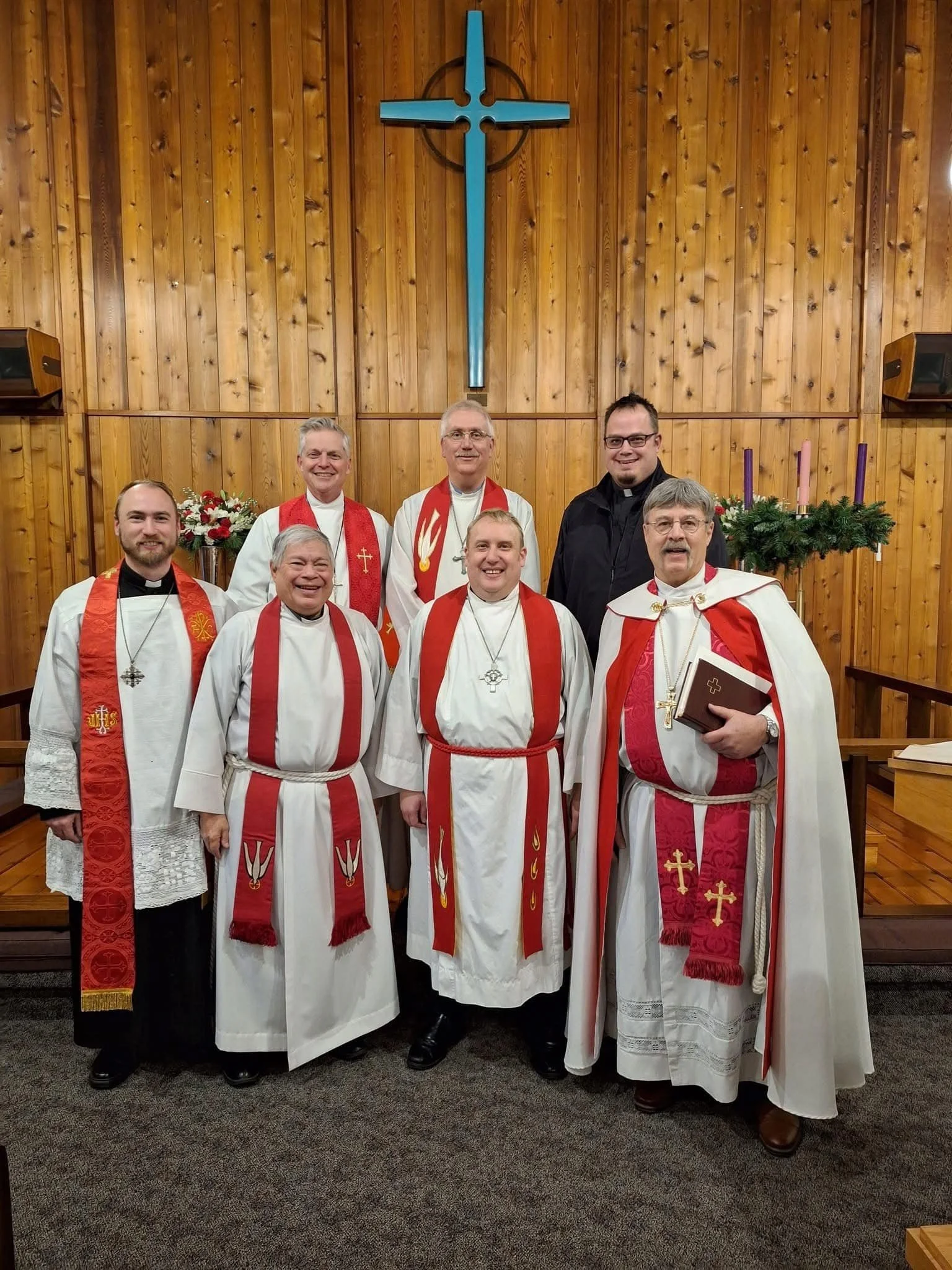 Seven men dressed in religious robes, posing inside a church with a wooden wall, a cross above them, and Christmas decorations, including an Advent wreath with candles and flowers.