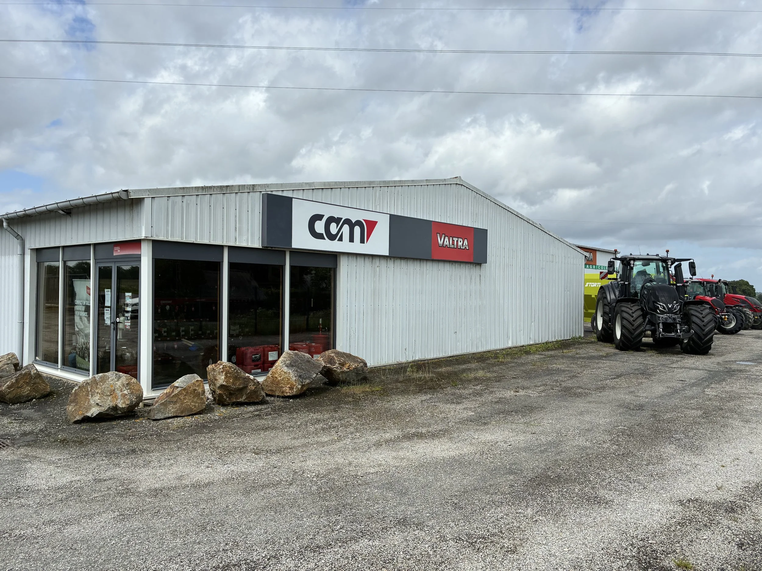 Un bâtiment de concession agricole avec le logo 'CAM' et 'Valtra' visible, entouré de plusieurs tracteurs agricoles noirs VALTRA.