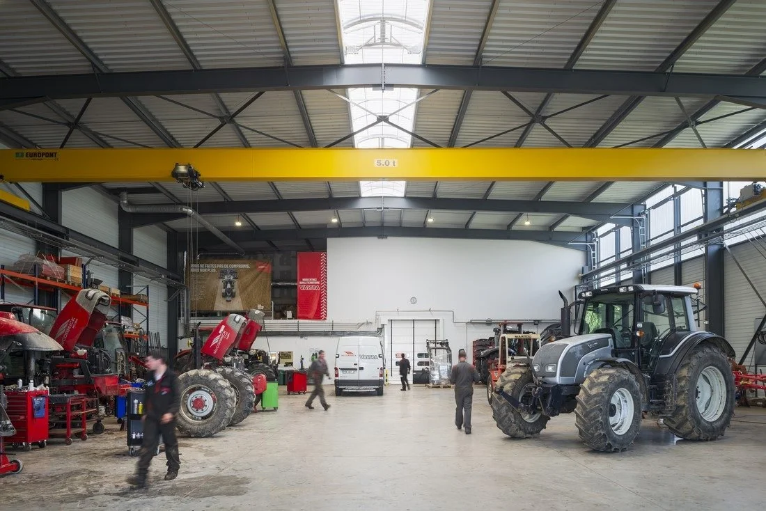 Un grand atelier de concession agricole avec plusieurs tracteurs agricoles et équipements en entretien ou répration. Pont roulant et équipement de pointe en second plan.