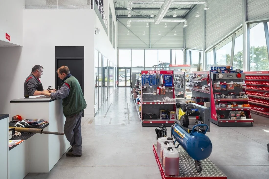 Intérieur d'un magasin libre-service agricole, avec un magasinier agricole au comptoir conseillant un client éleveur avec rayons d'outillage en arrière-plan.