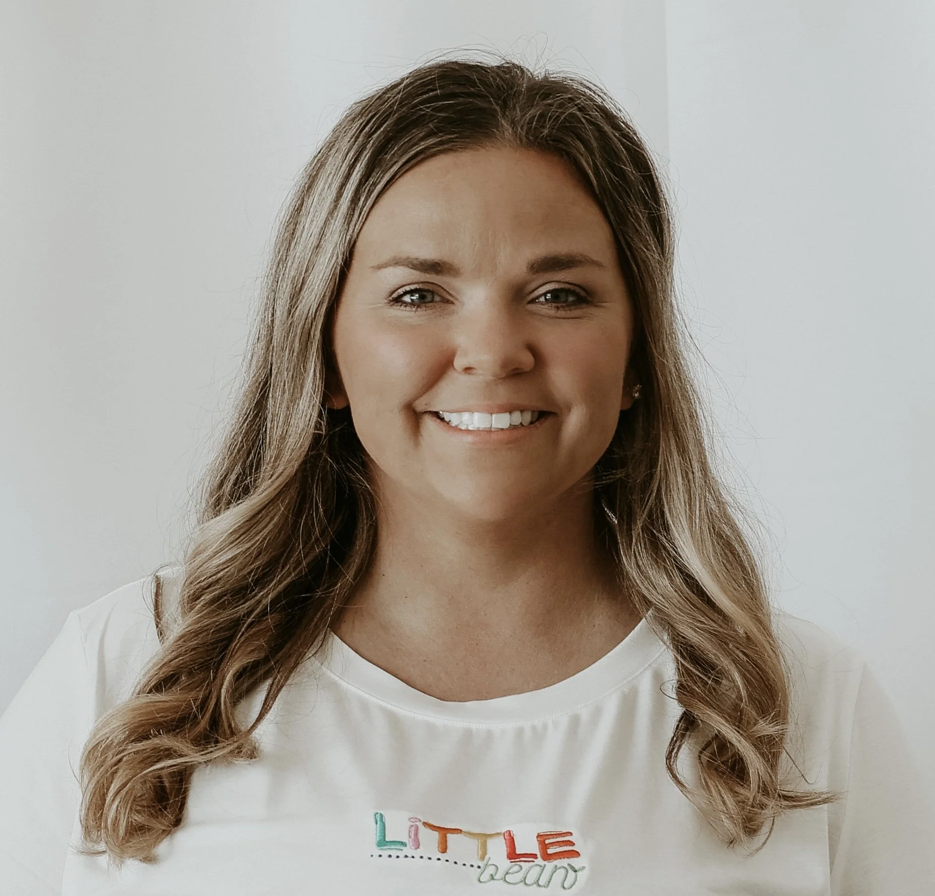 Close-up of a woman with long, wavy brown hair smiling against a plain light background, wearing a white top with colorful text.