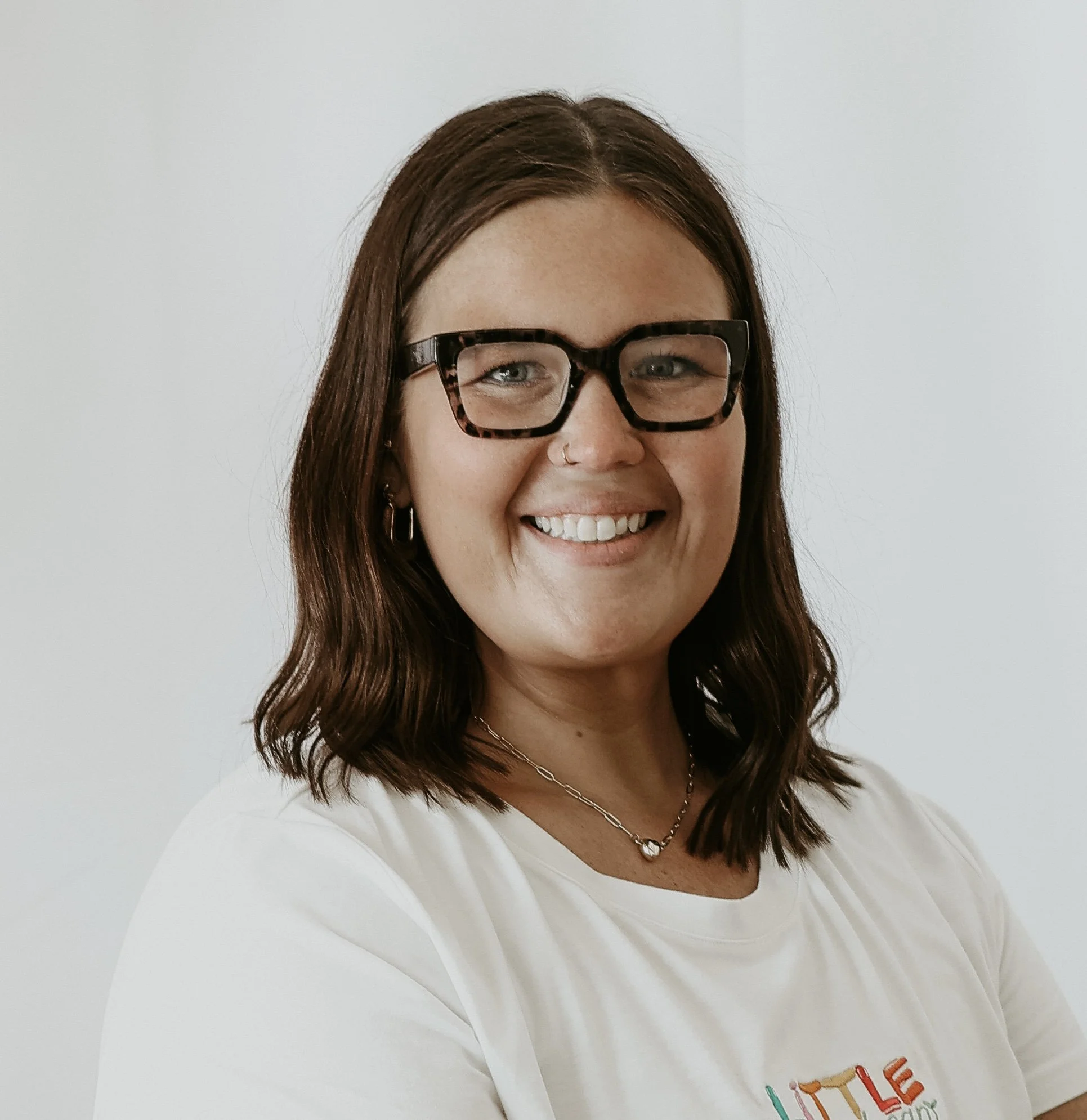 Close-up of a woman with shoulder-length brown hair, wearing tortoise-shell glasses, a nose ring, and a necklace, smiling in front of a plain light-colored wall.