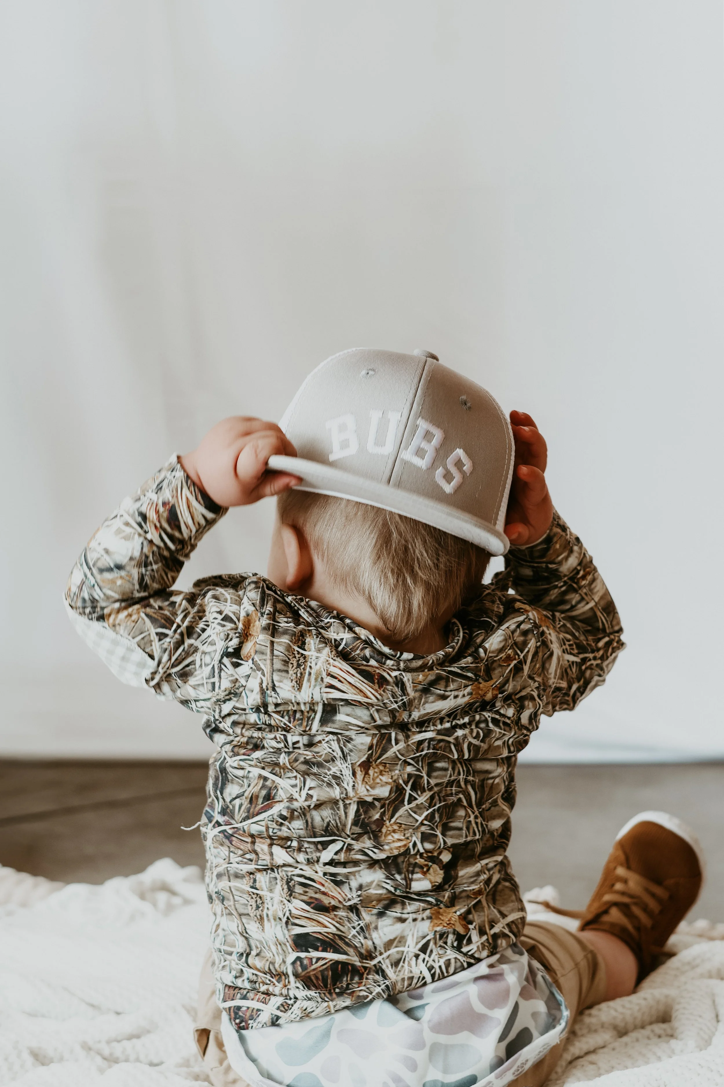 A young child kneeling on a fluffy white rug, wearing a camouflage patterned long-sleeve shirt, beige shorts with a camouflage print, and a beige baseball cap with the word 'BUBS' on it. The child is adjusting the cap, facing away from the camera, with a white wall in the background.