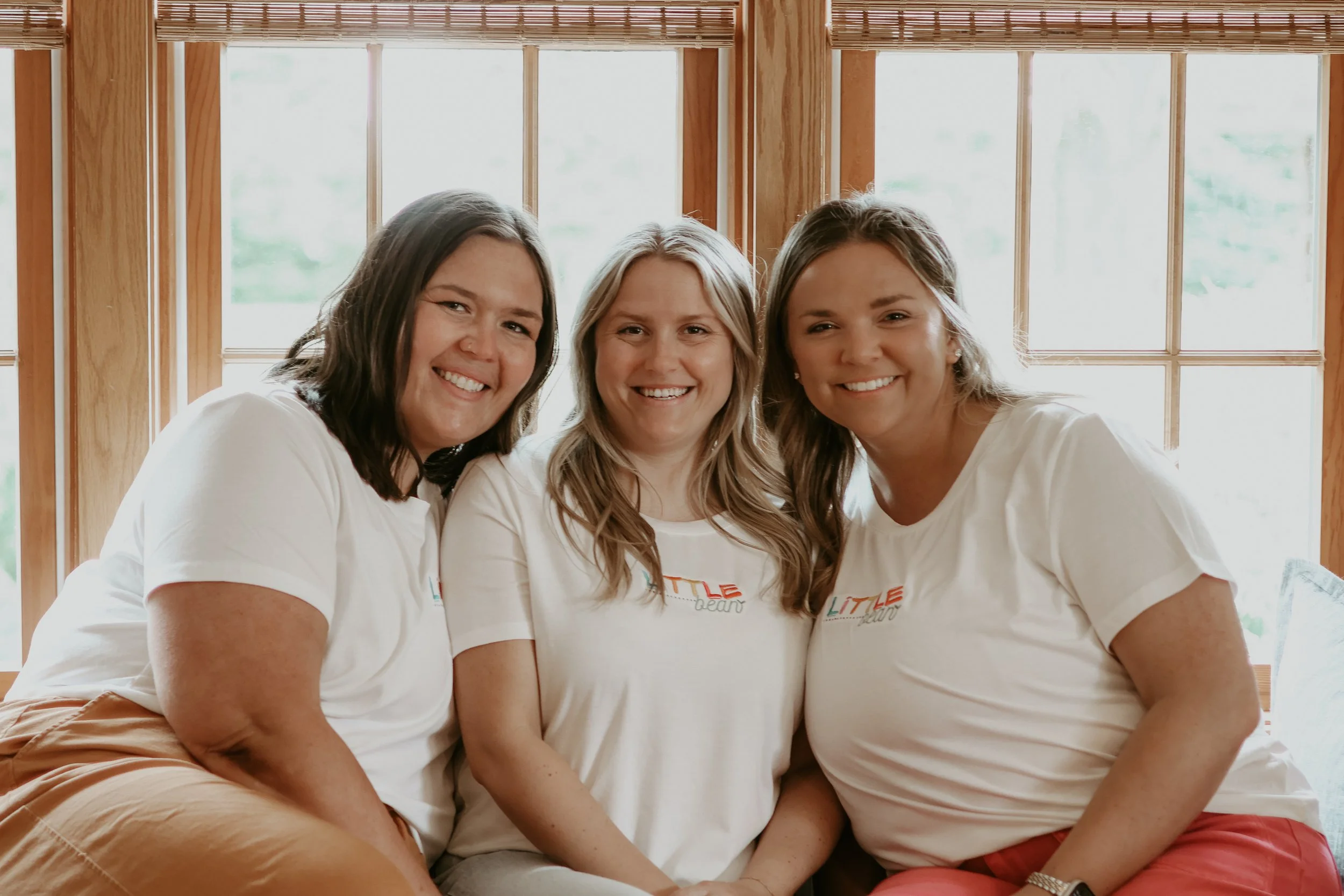 Three women sitting close together indoors near large windows, smiling at the camera. They are wearing white t-shirts.