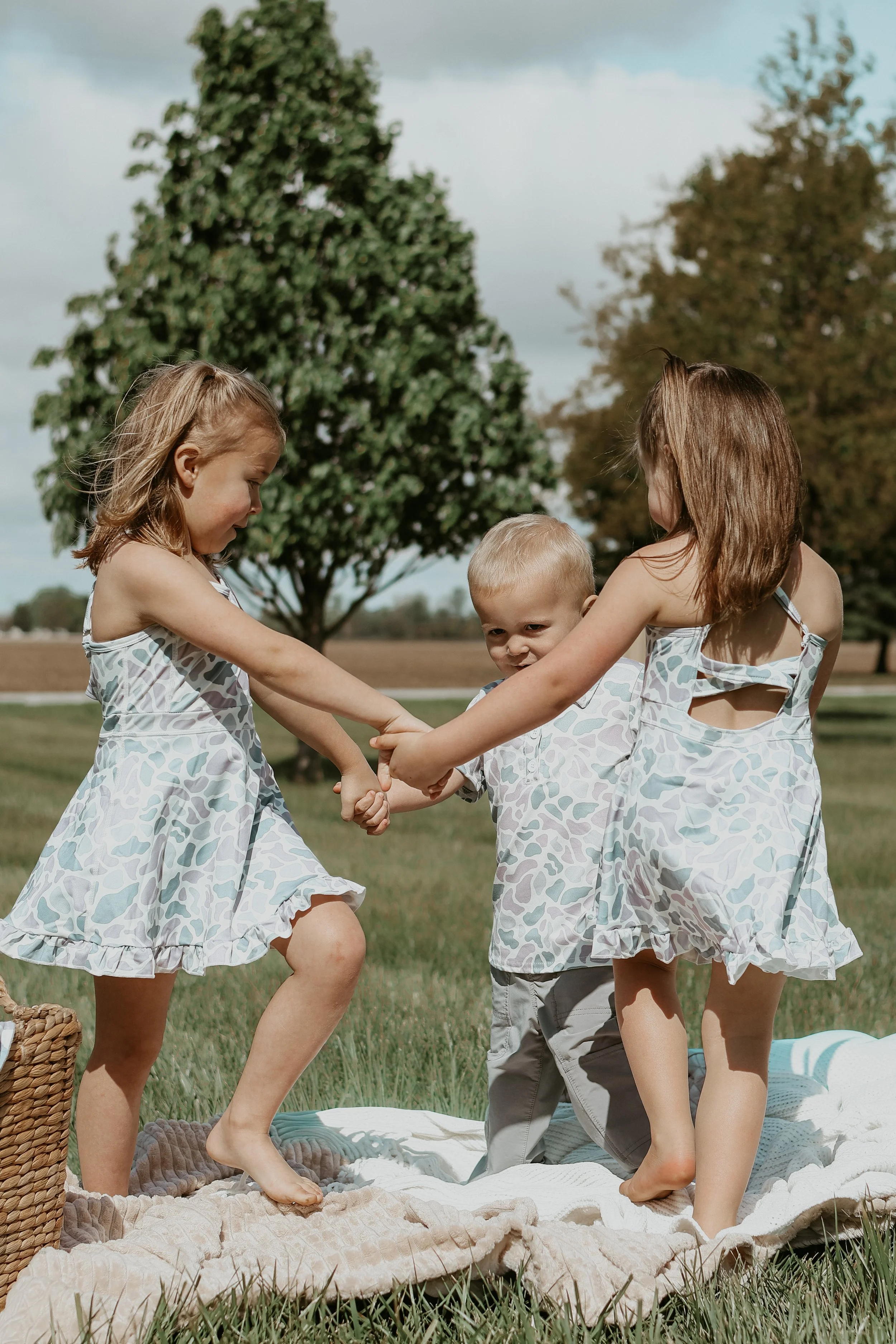 Four children, two girls and two boys, hold hands and play together outside on a blanket in a park with trees and cloudy sky in the background.