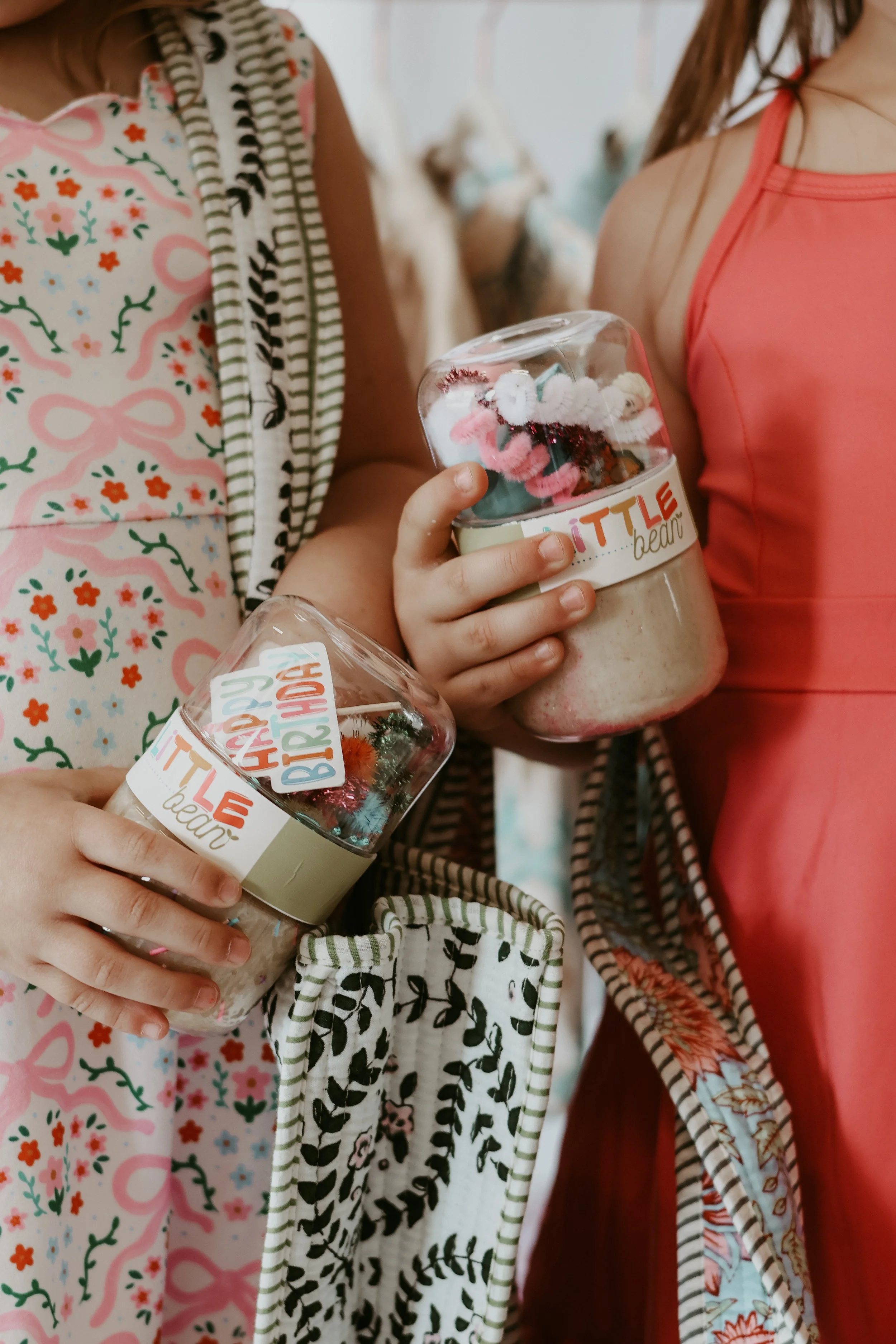 Close-up of two children holding jars filled with craft supplies, such as yarn and decorations, with colorful patterned clothing and scarves.