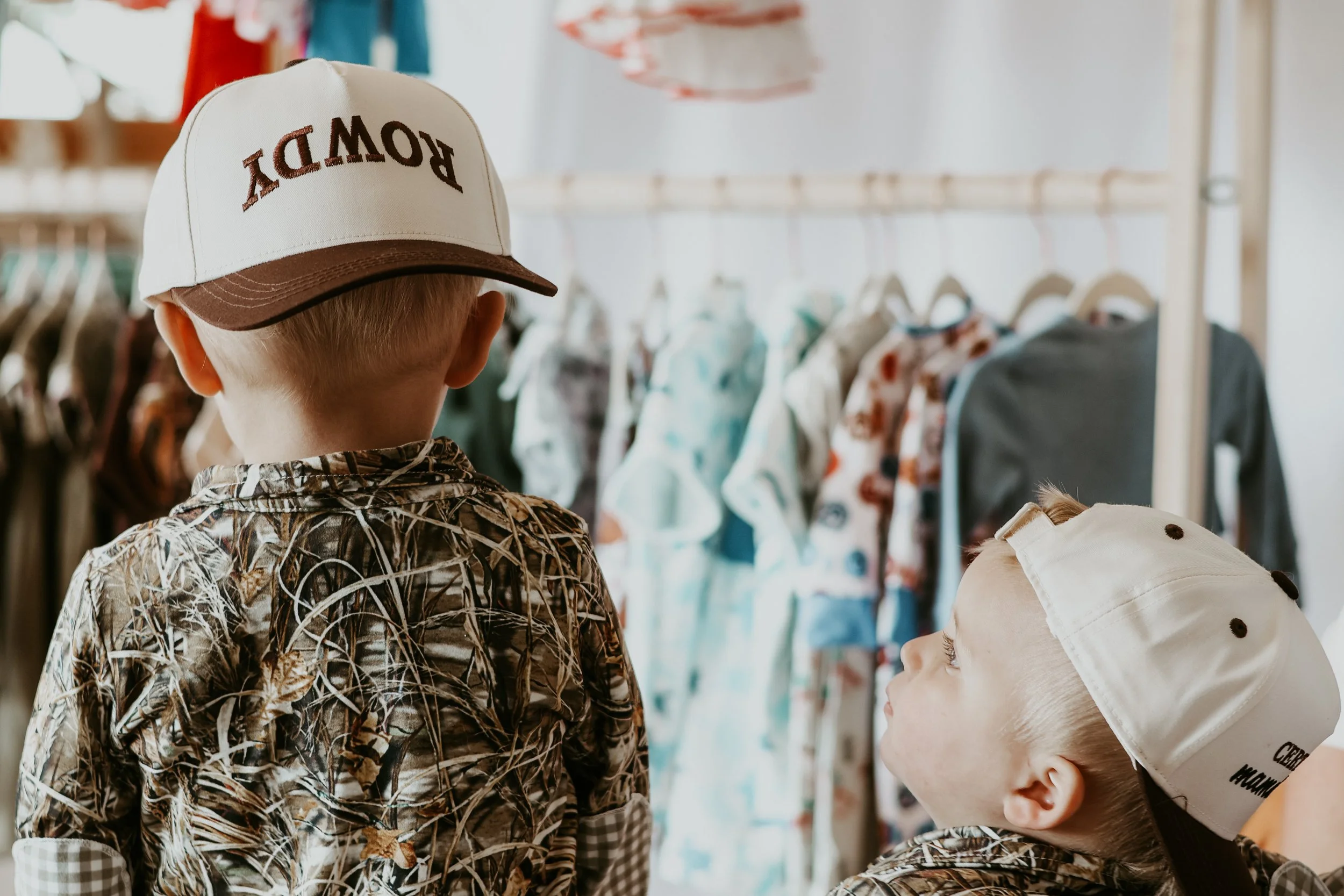 Two young children, one boy and one girl, shopping for clothes in a store. The boy is wearing a palm print shirt and a beige cap with the word "ROWDY" on it, while the girl has on a matching palm print shirt and a beige hat with black polka dots, with clothing racks filled with colorful garments in the background.