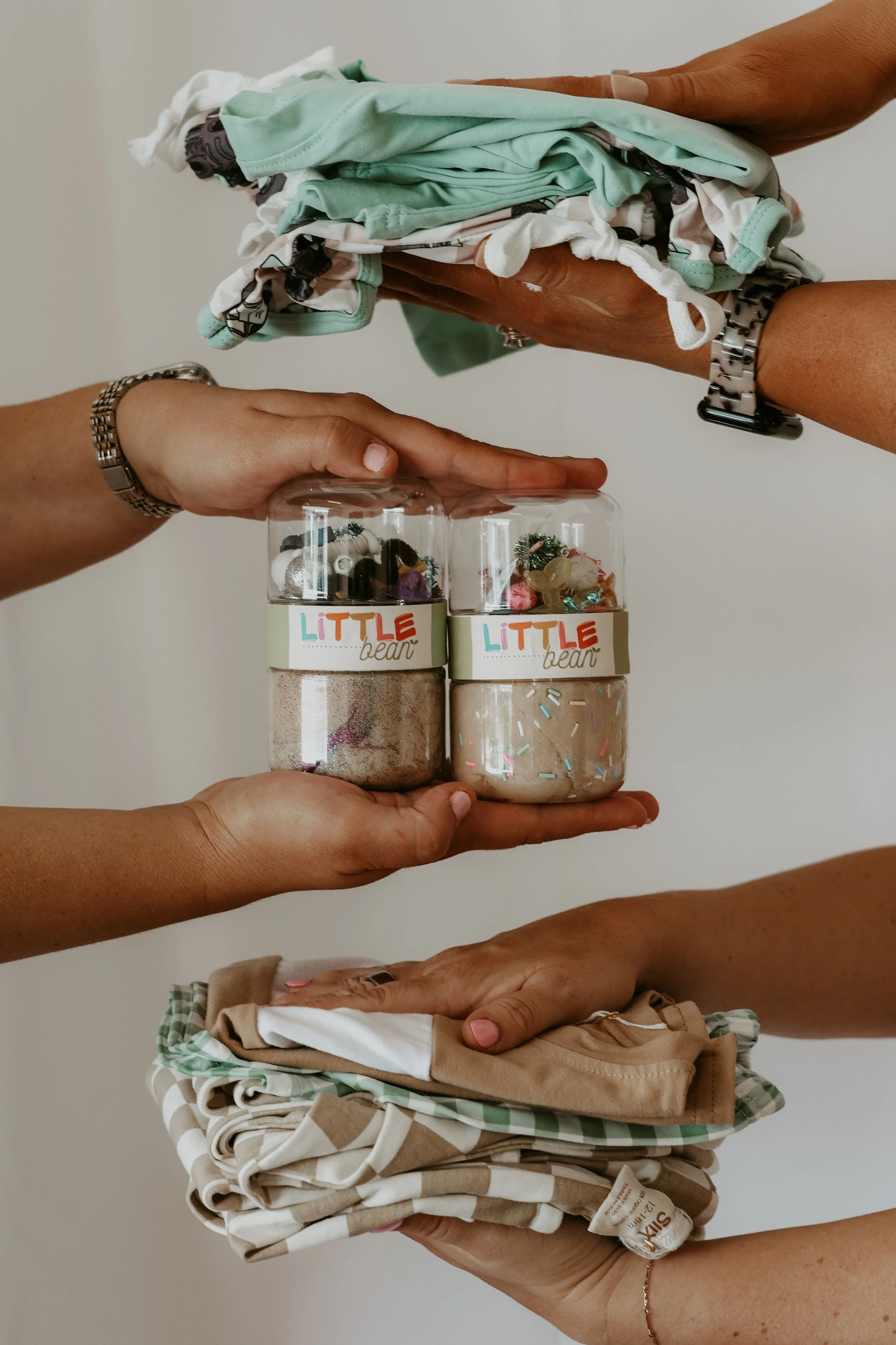 Multiple hands holding piles of clothing and clear jars filled with small objects. The jars are labeled 'Little Bear.'
