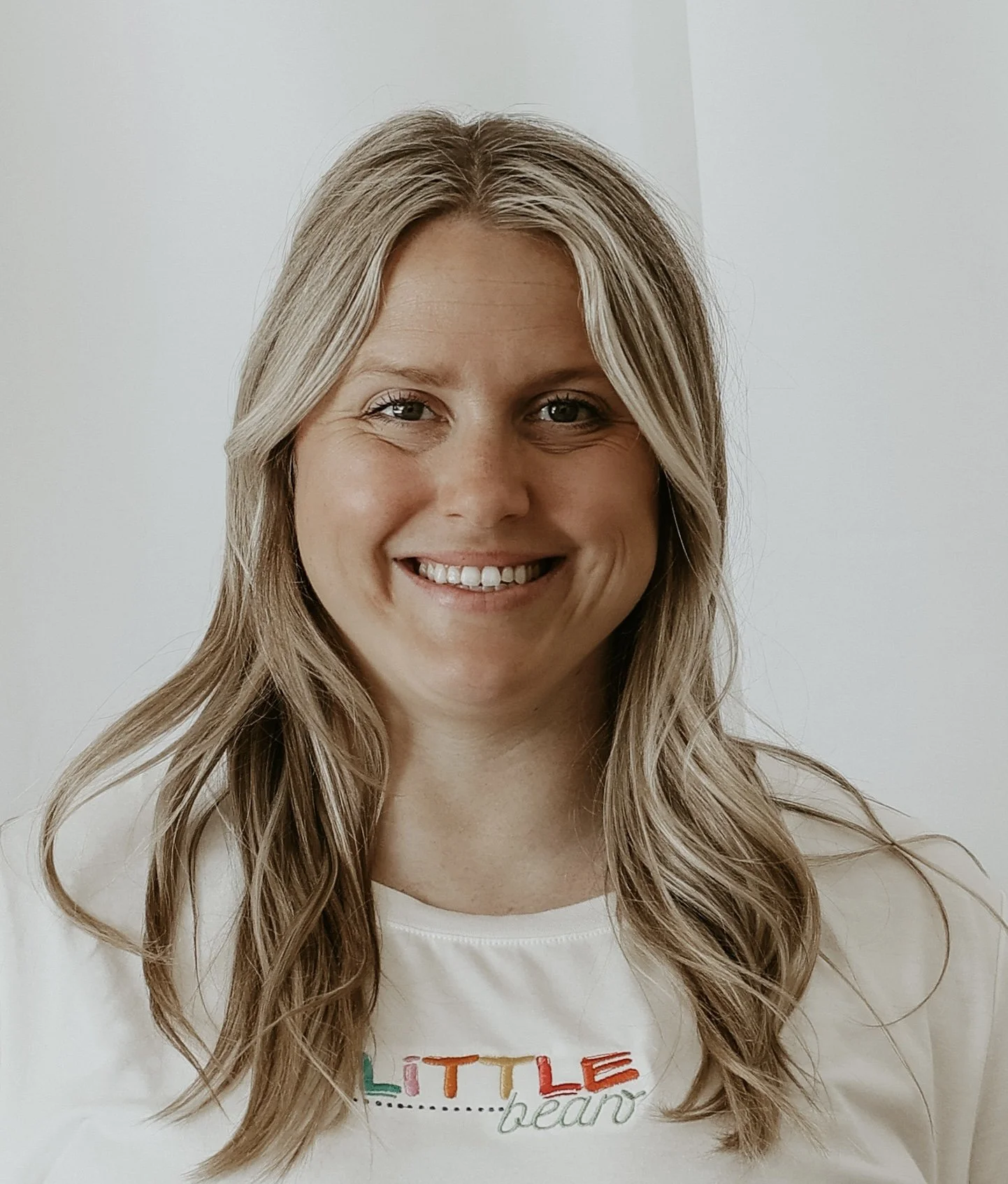 A woman with long, wavy blonde hair smiling against a plain background, wearing a white t-shirt with colorful text.