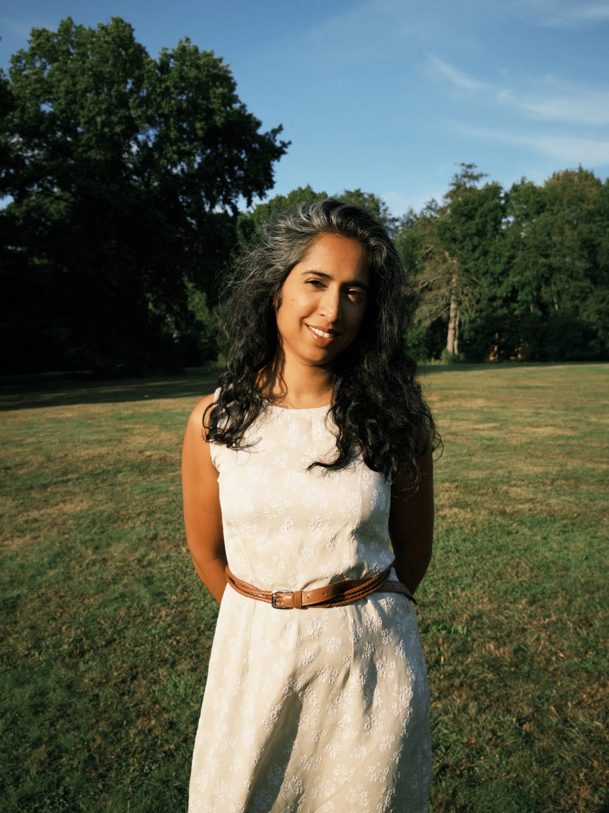A woman with long curly dark hair standing in a grassy park with trees and blue sky in the background.