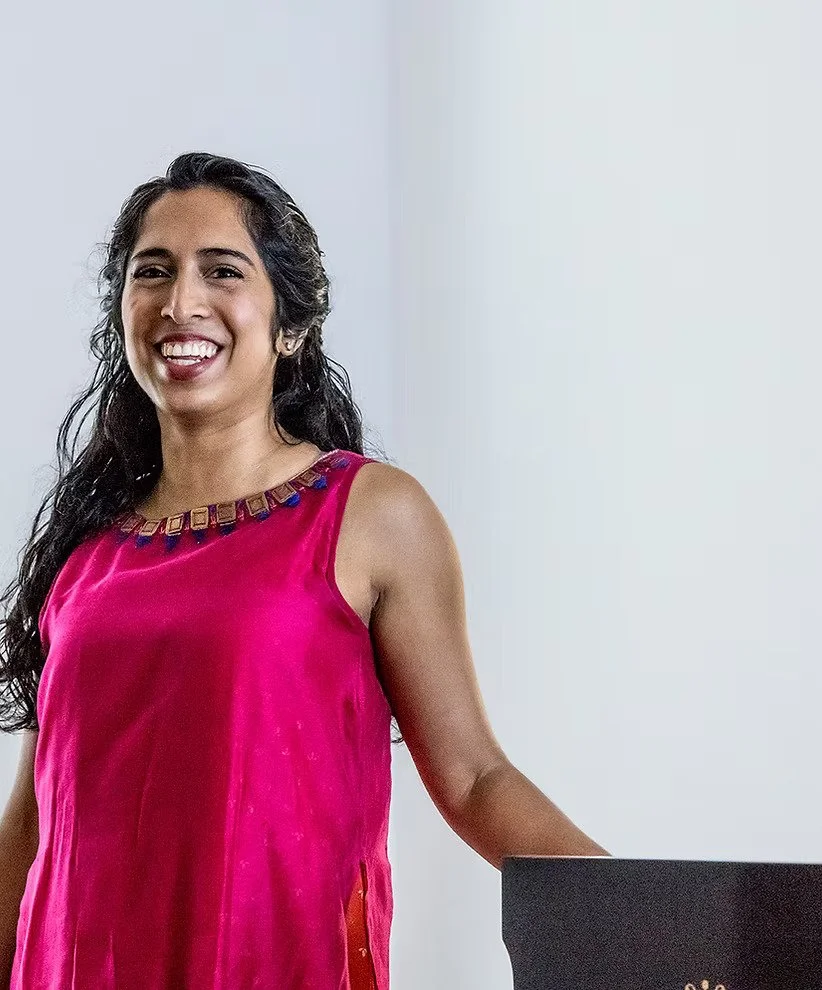 A woman smiling with long dark hair, wearing a sleeveless pink top with decorative accents around the neckline, standing next to a black object against a plain gray and white background.