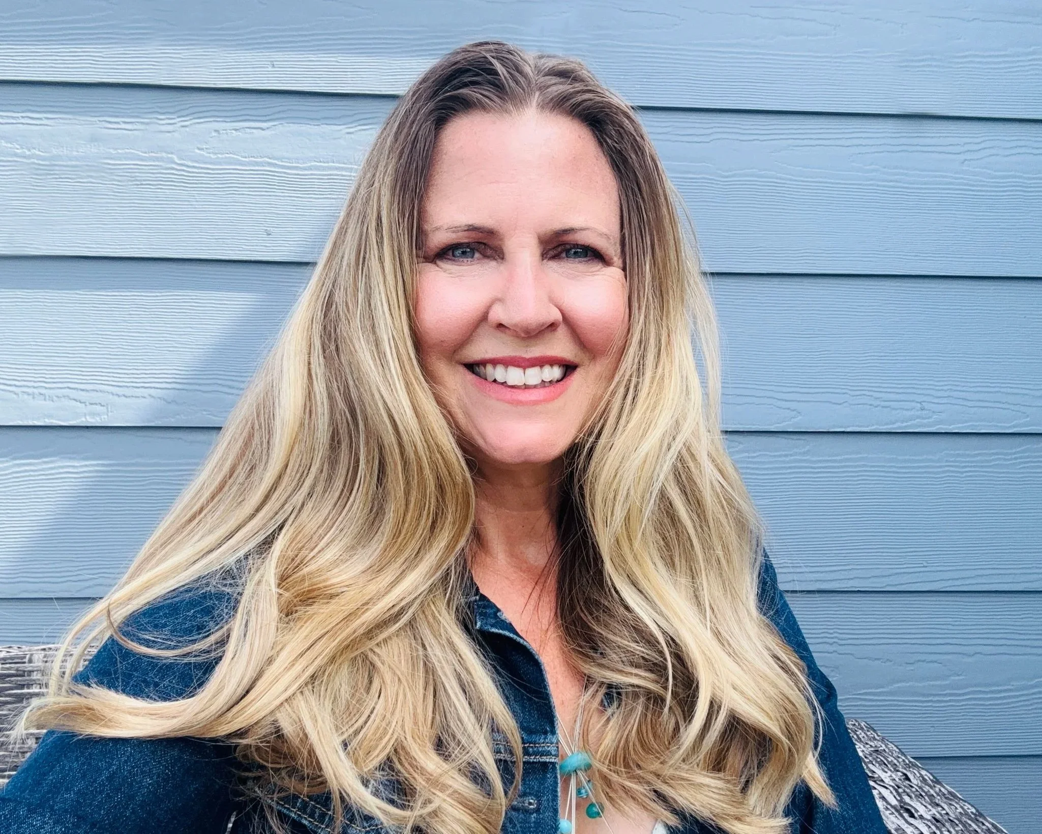 Portrait of a smiling woman with long blonde hair standing in front of light blue horizontal wooden siding.