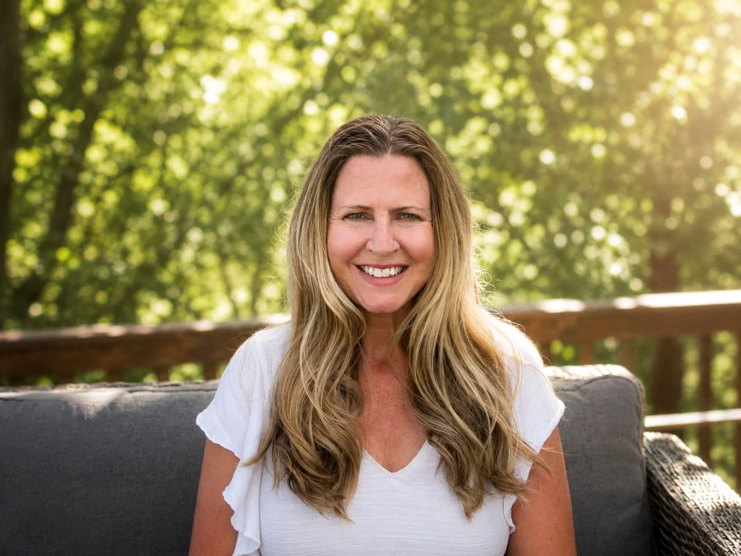 A smiling woman with long, wavy blonde hair sitting outdoors on a lounge chair with a wooden railing and lush green trees in the background.