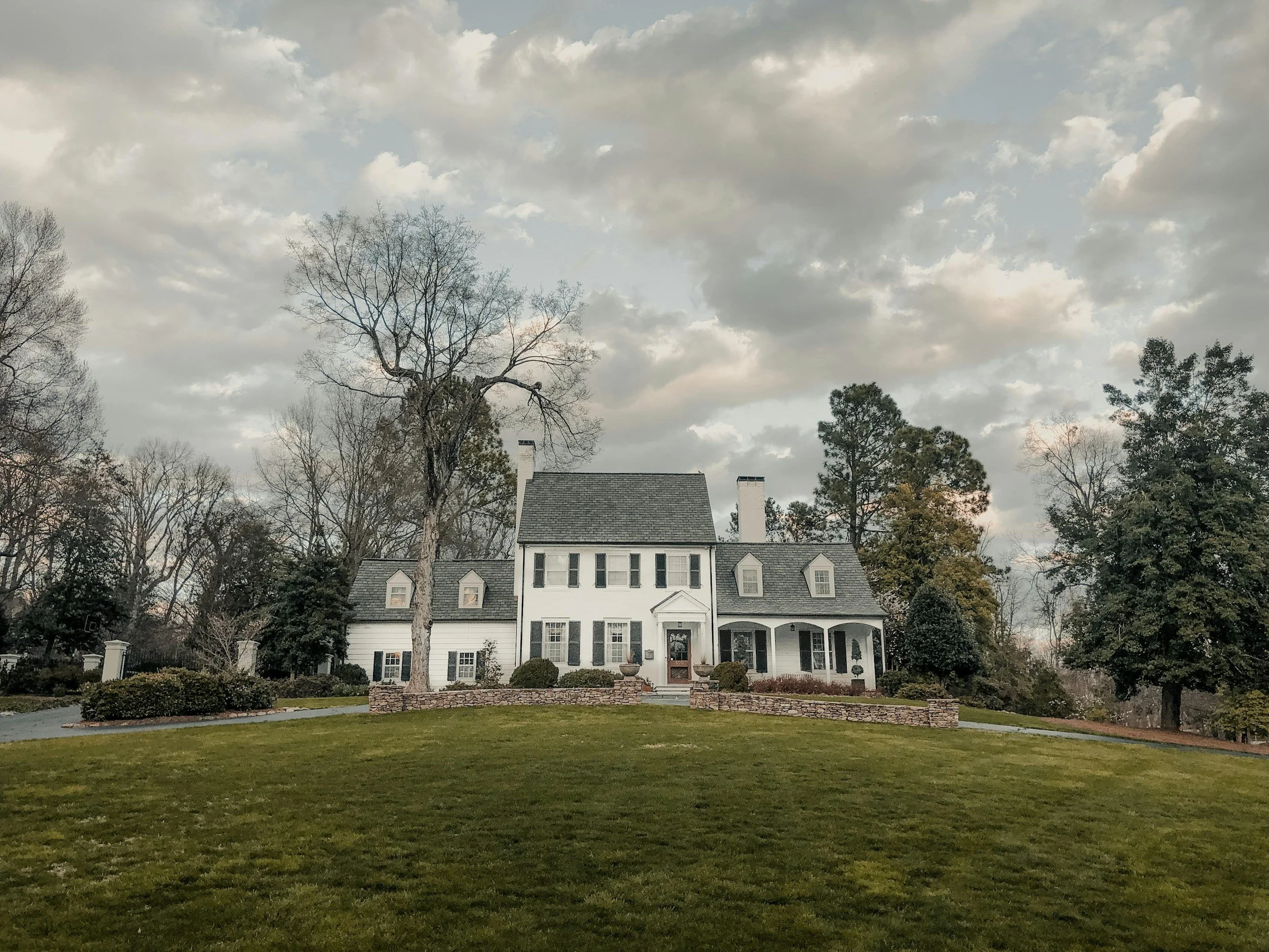 Large, white, two-story house with black shutters and a gray roof, surrounded by a well-maintained lawn and trees under a cloudy sky.