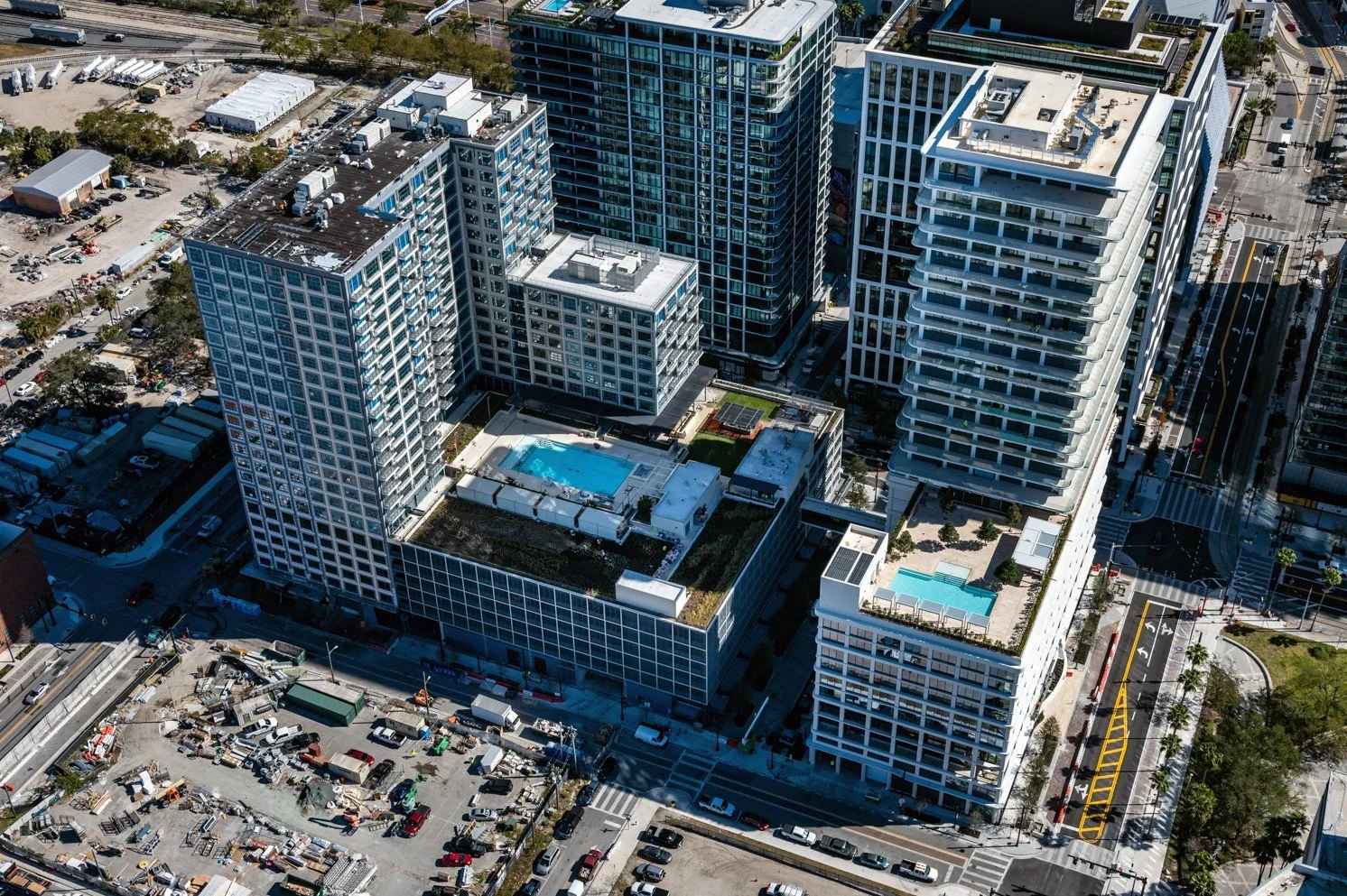 High-angle aerial view of modern skyscrapers with rooftop swimming pools, surrounded by city streets, intersecting roads, and nearby construction area with parked vehicles.