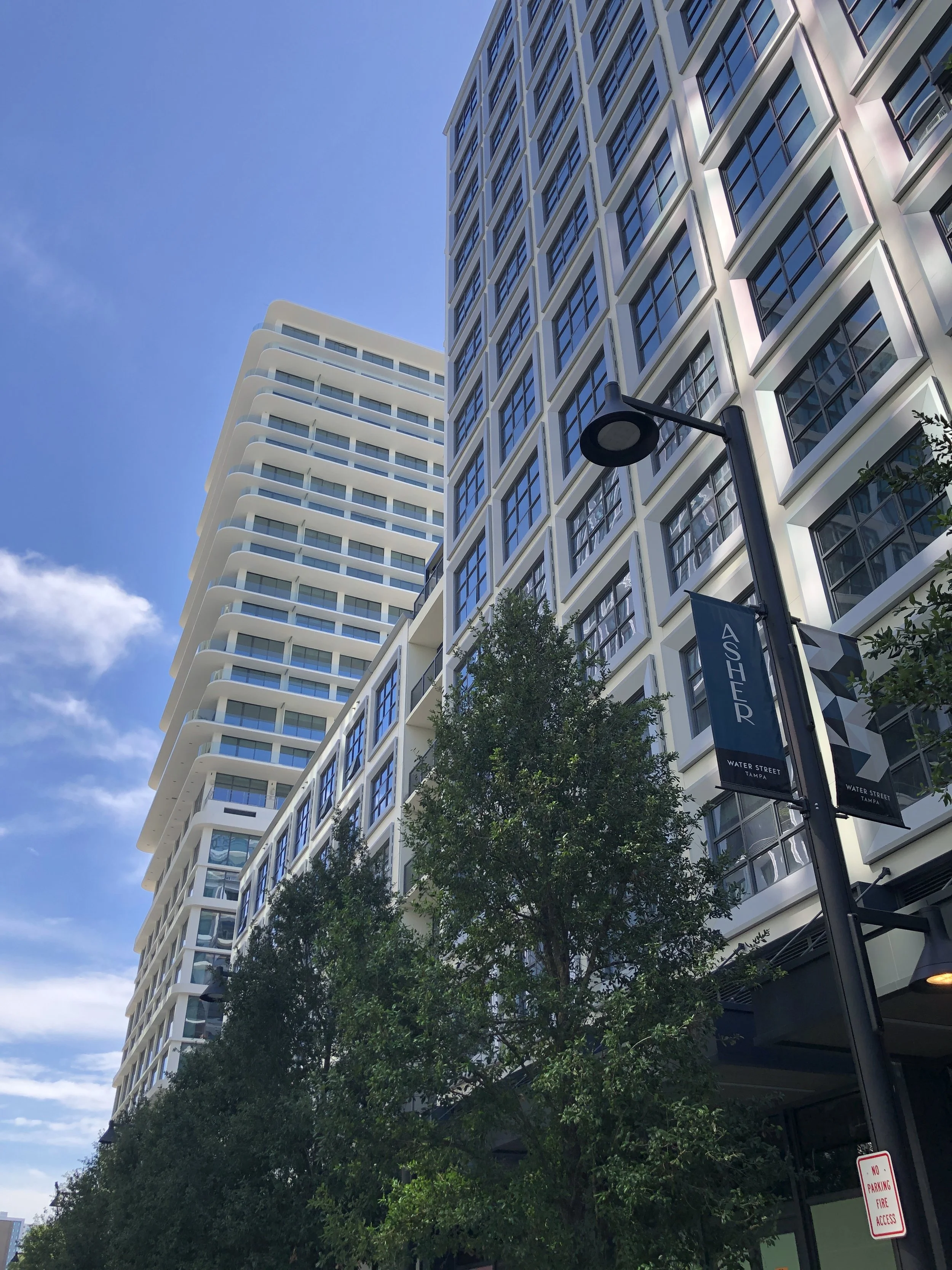 Tall modern building with numerous windows, a street lamp, trees, a blue sky, and banners for Water Street Tampa.