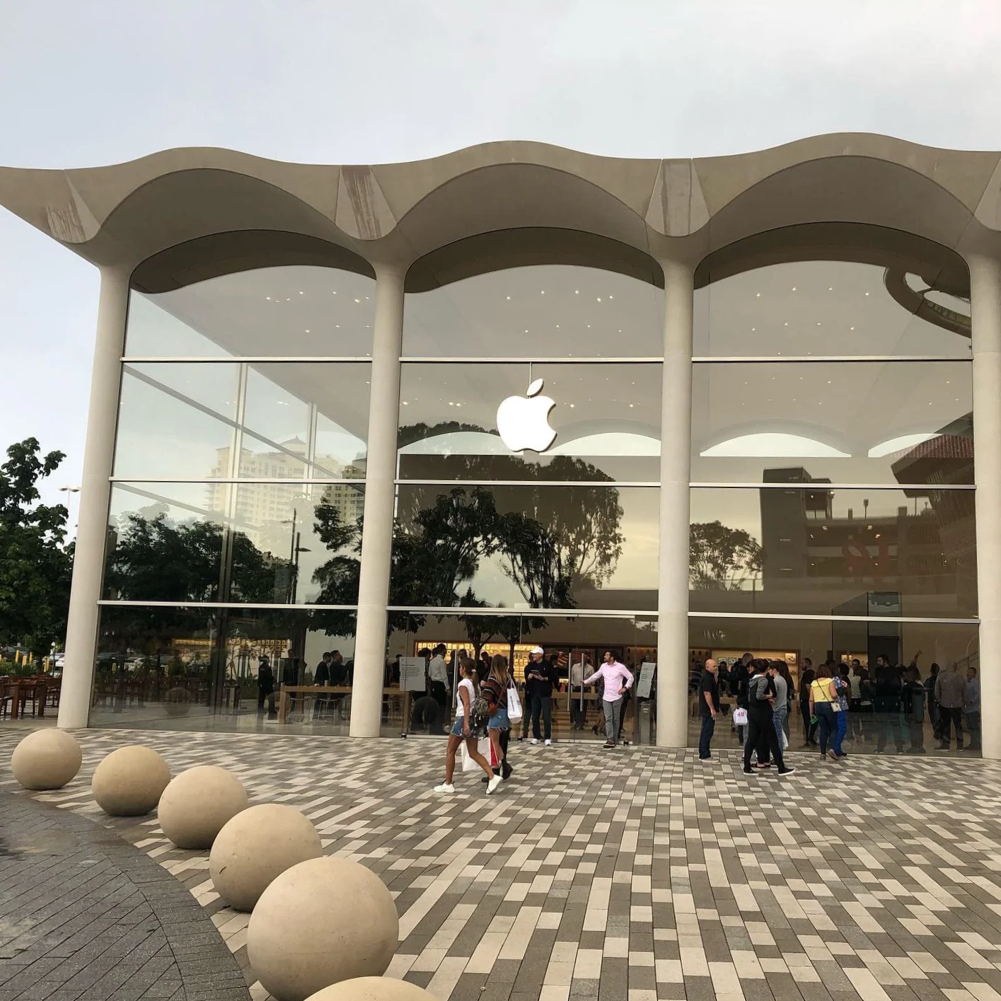 Apple Store with large glass facade and white logo, shoppers entering and exiting, patterned pavement, spherical stone decorations outside.