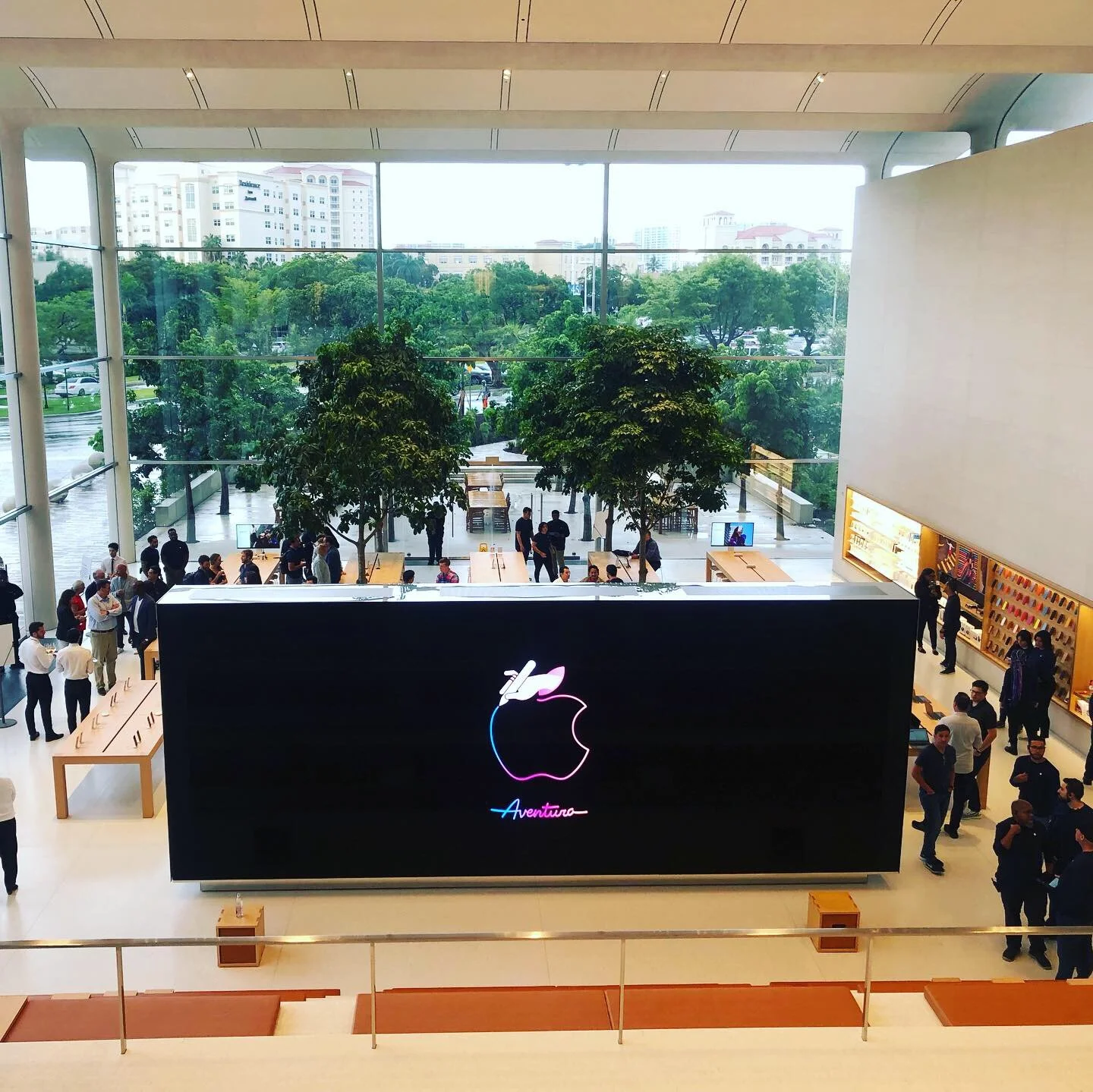 Inside an Apple Store with a large black sign featuring the colorful Apple logo and the word 'Aventura' in neon, people shopping and interacting, trees visible through large glass windows, and a display with Apple products on the right.