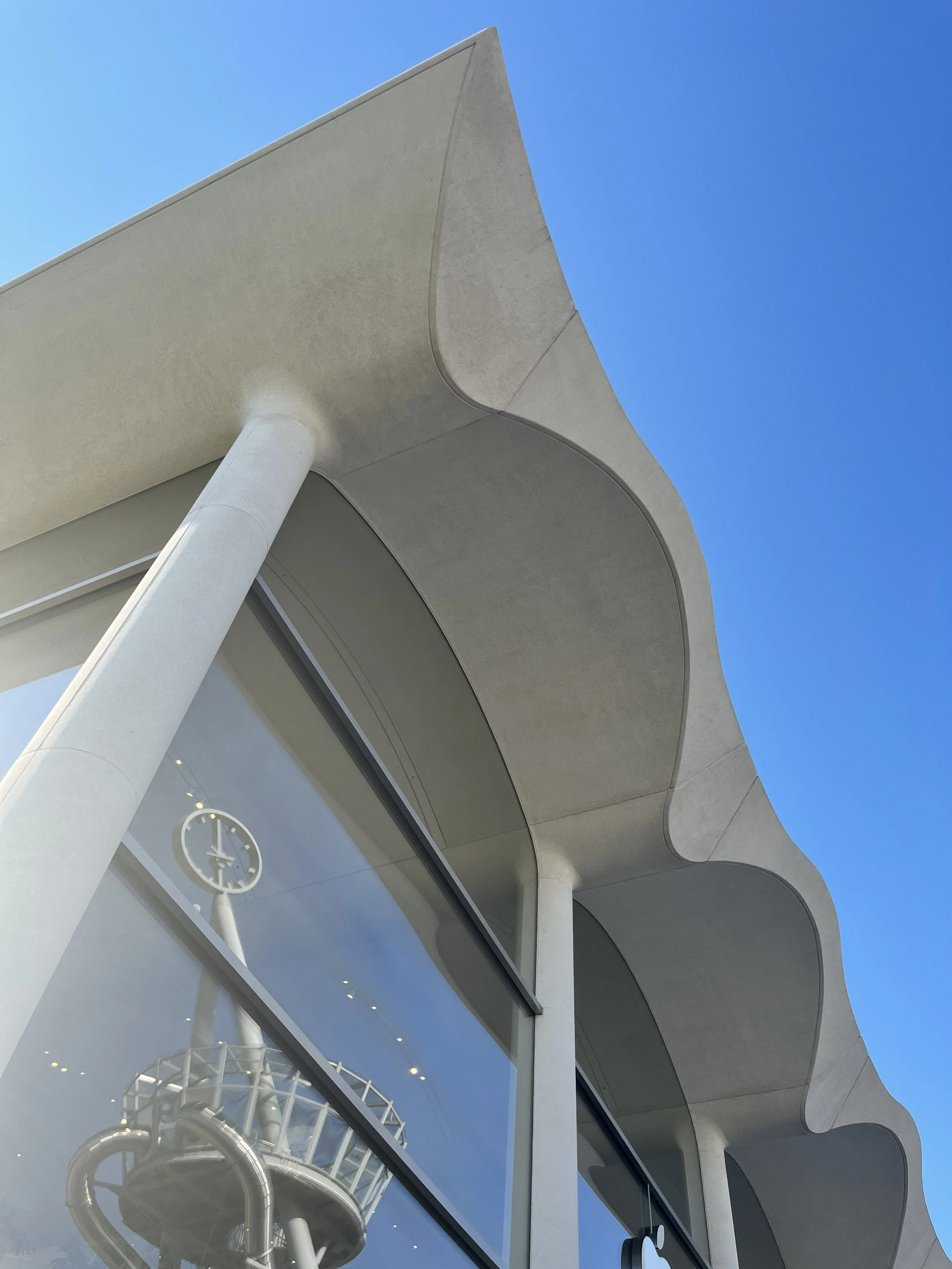 Looking up at a modern building with curved architectural design, large glass windows, and a clock visible through the glass, with a clear blue sky background.