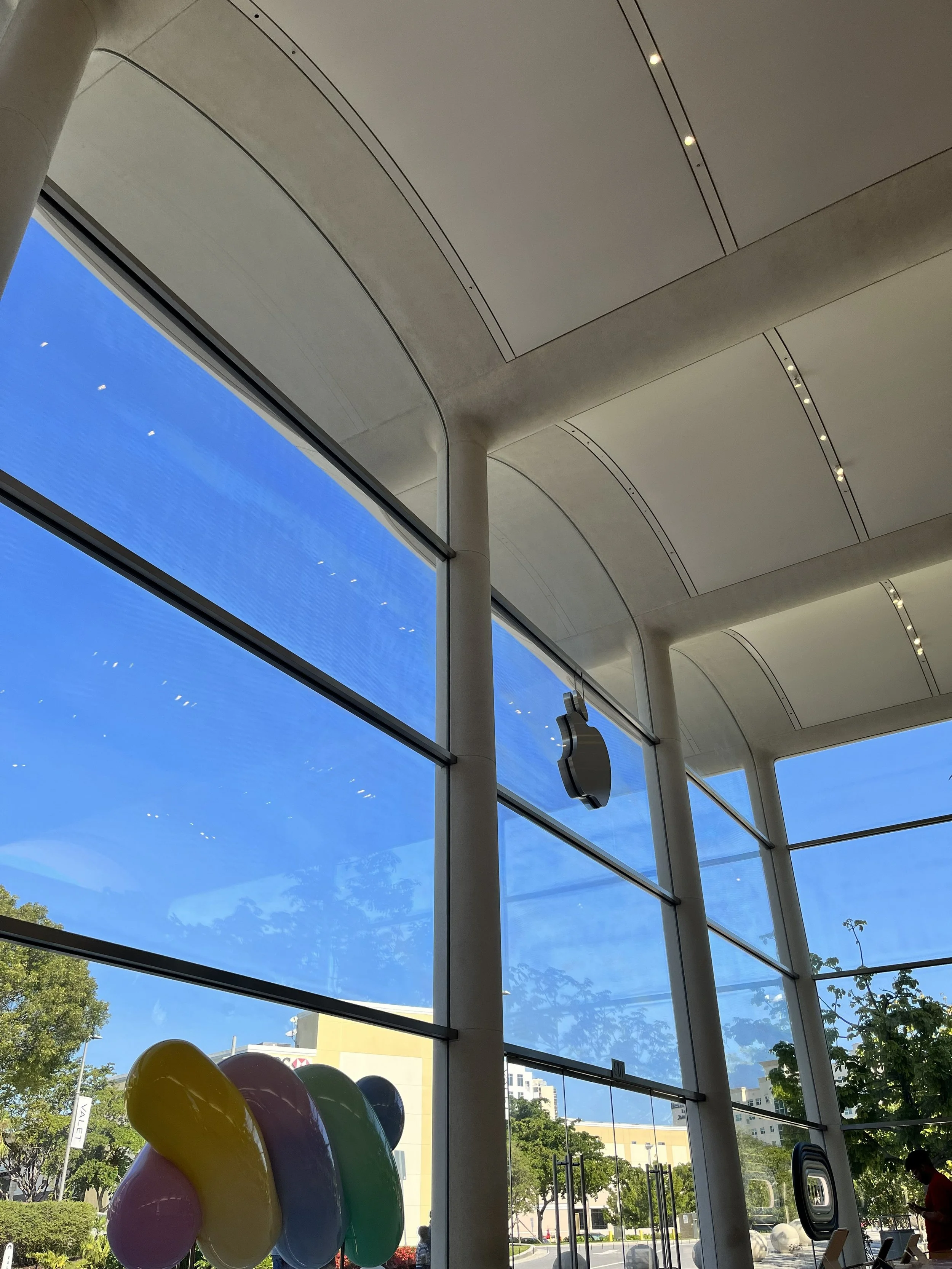 Interior view of the Apple Store with large windows showing a blue sky and trees outside. Colorful balloons are displayed near the window, and the store features modern architecture with curved ceilings and ceiling lights.