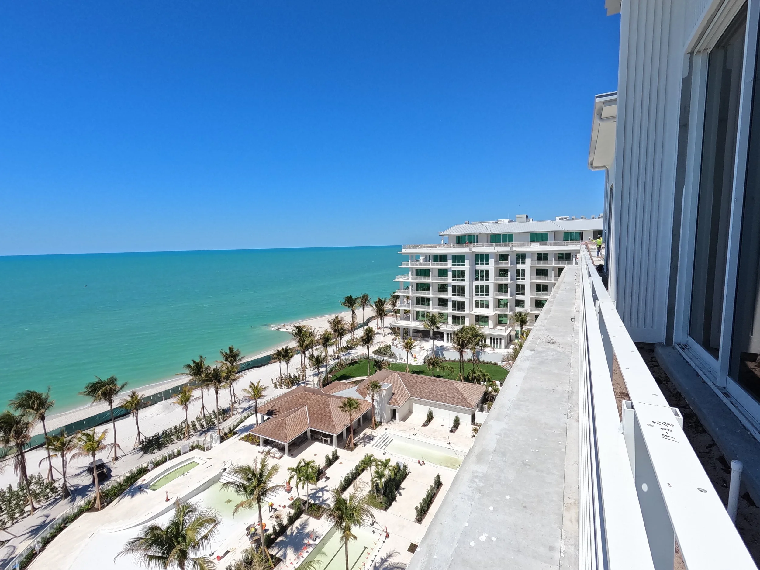 View from a high-rise balcony overlooking a beach with turquoise water, palm trees, and a resort with multiple floors and balconies under a clear blue sky.