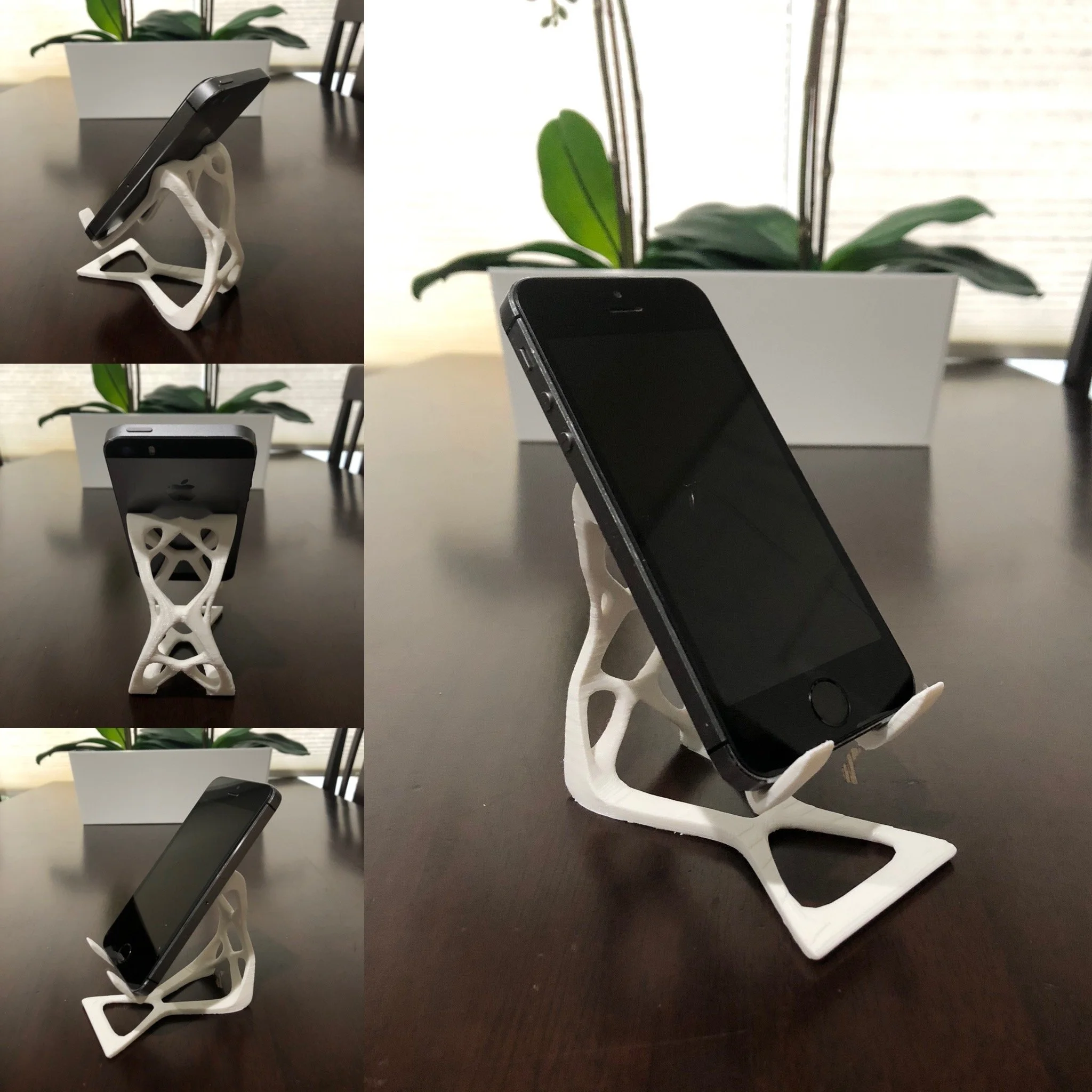 A black smartphone resting on a white decorative stand on a dark brown table. A potted plant is visible in the background.