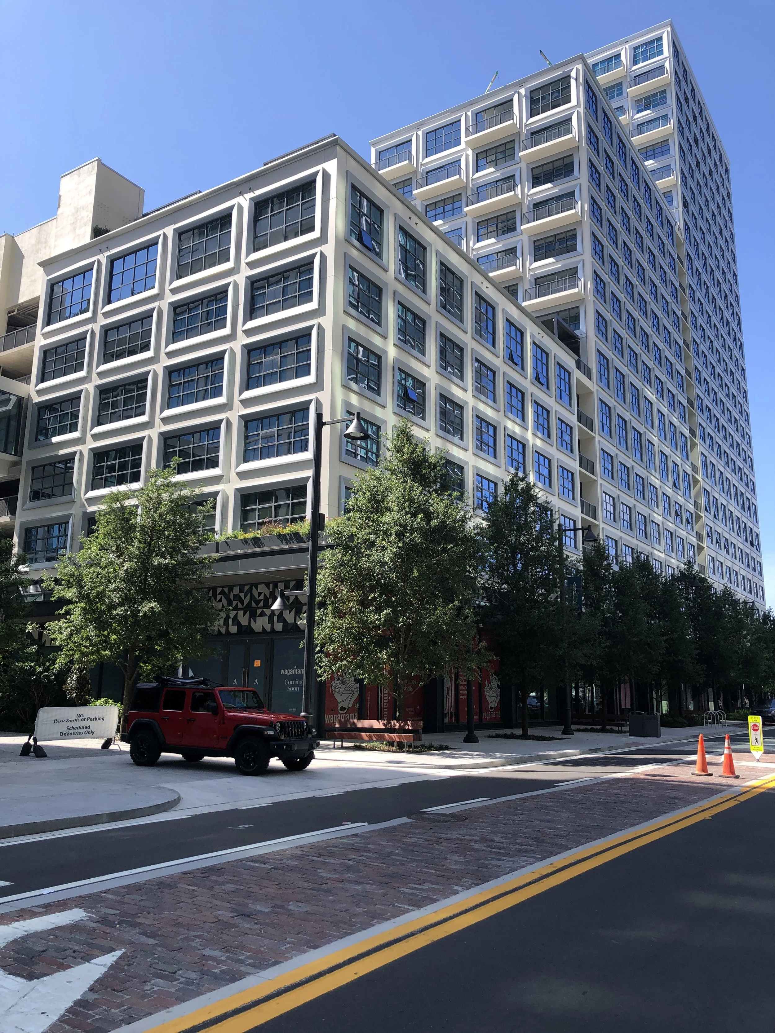 A city street with a modern multi-story white building with large windows, a red Jeep parked on the side, trees lining the sidewalk, and orange traffic cones near the curb. Clear blue sky overhead.