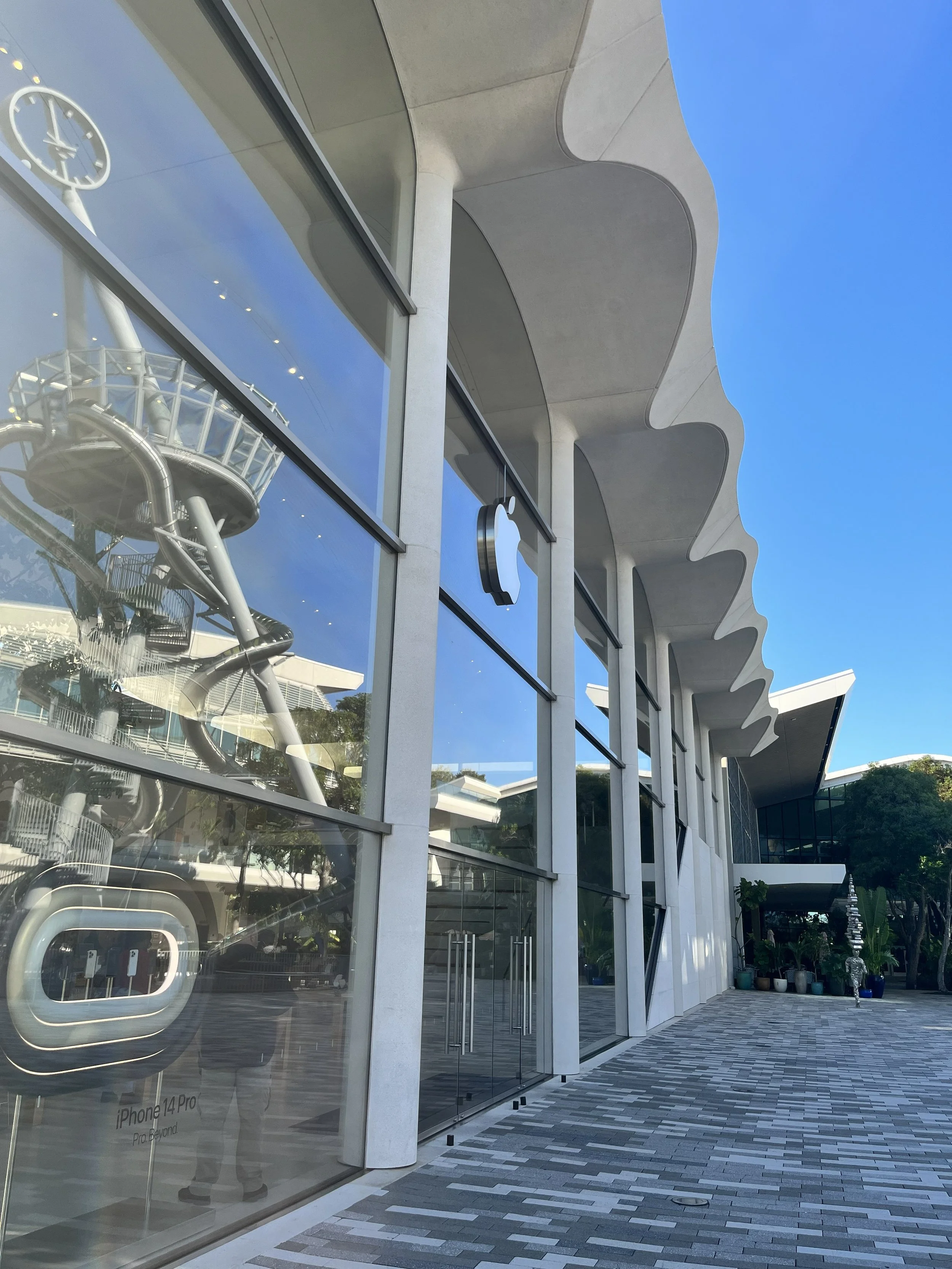 Exterior view of an Apple Store with large glass windows and a white wavy architectural roof, reflecting the ferris wheel and blue sky, with potted plants and a person walking outside.