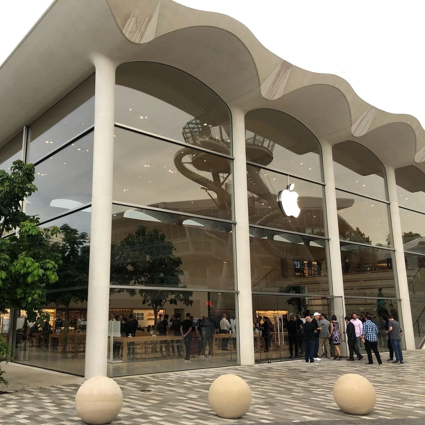 Exterior view of an Apple Store with large glass windows and a visible spiral staircase inside; people are gathered outside the store entrance.