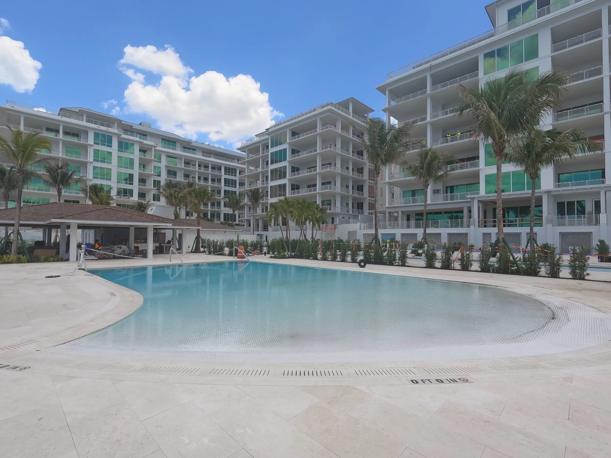 Outdoor pool area in front of modern apartment buildings with palm trees and a clear blue sky.