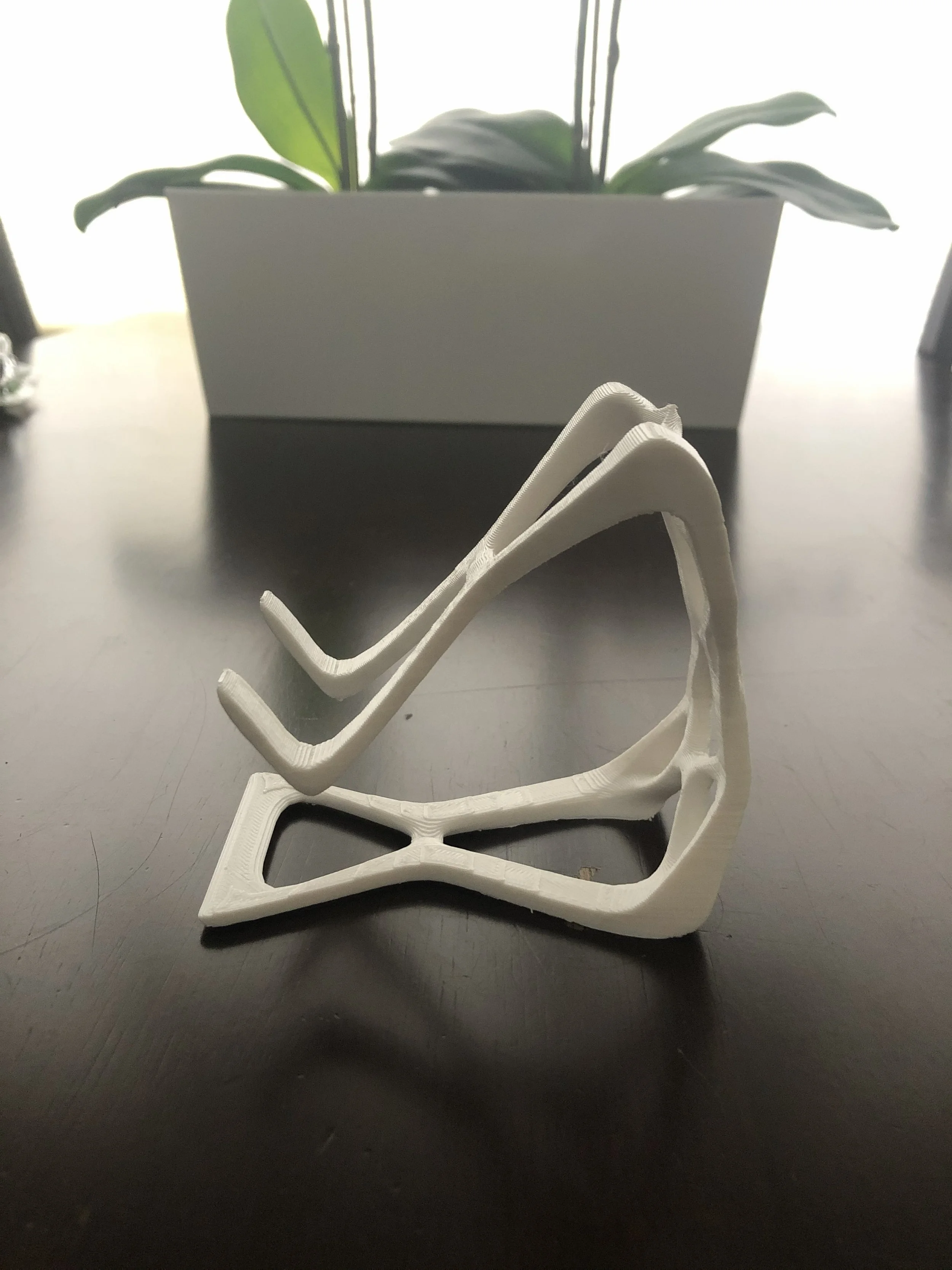 A white 3D-printed chair model placed on a dark surface with a blurred plant and white background in the back.