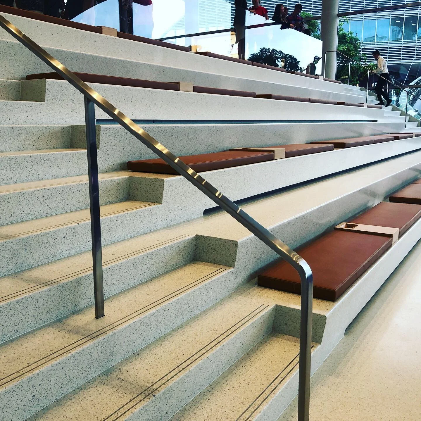 Indoor stairway with terrazzo steps, brown cushioned seats, and a metal handrail, with people standing at the top and walking down.