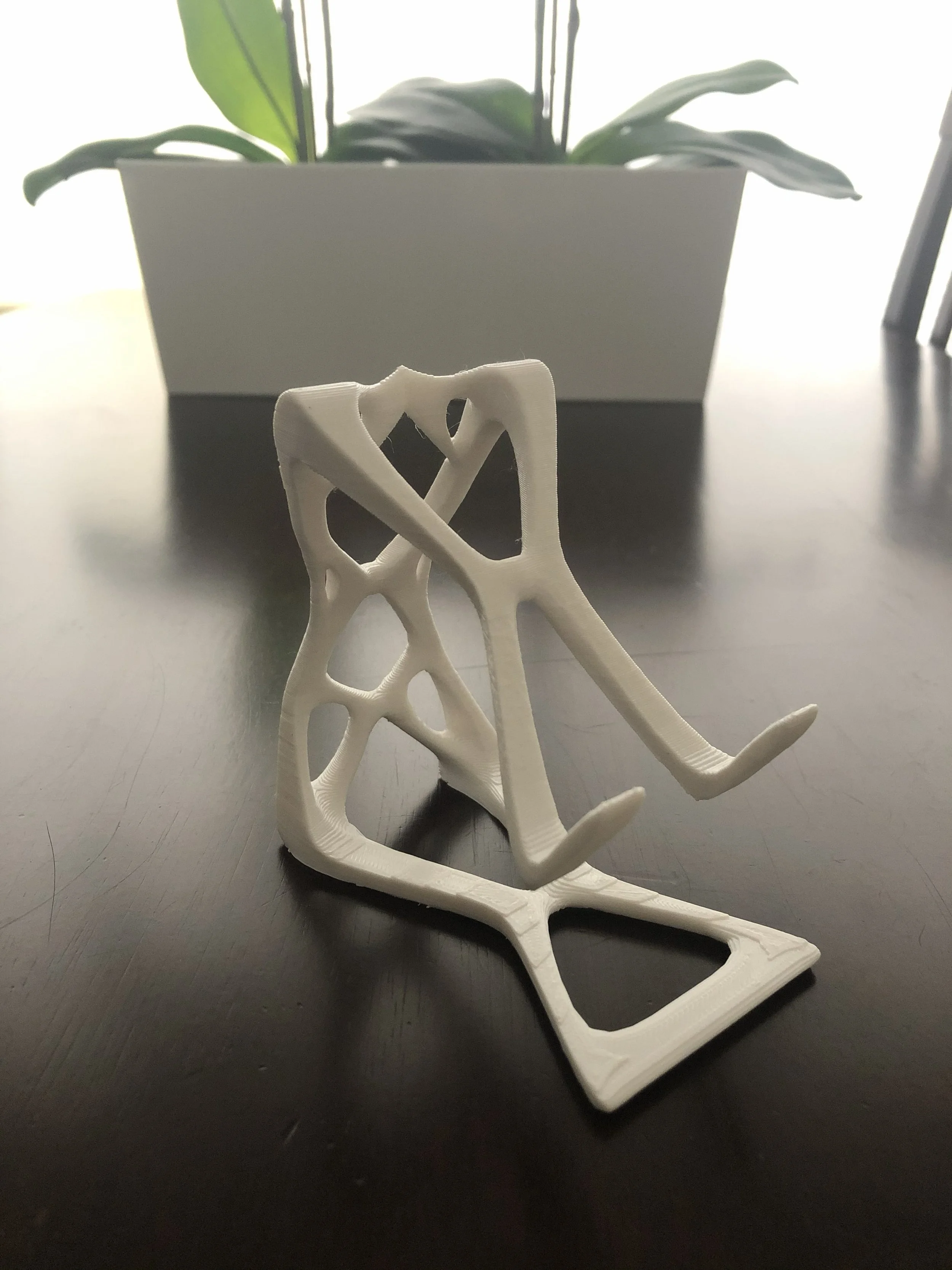 A white 3D-printed sculpture of a human skull, placed on a black surface with a plant in a rectangular pot in the background.