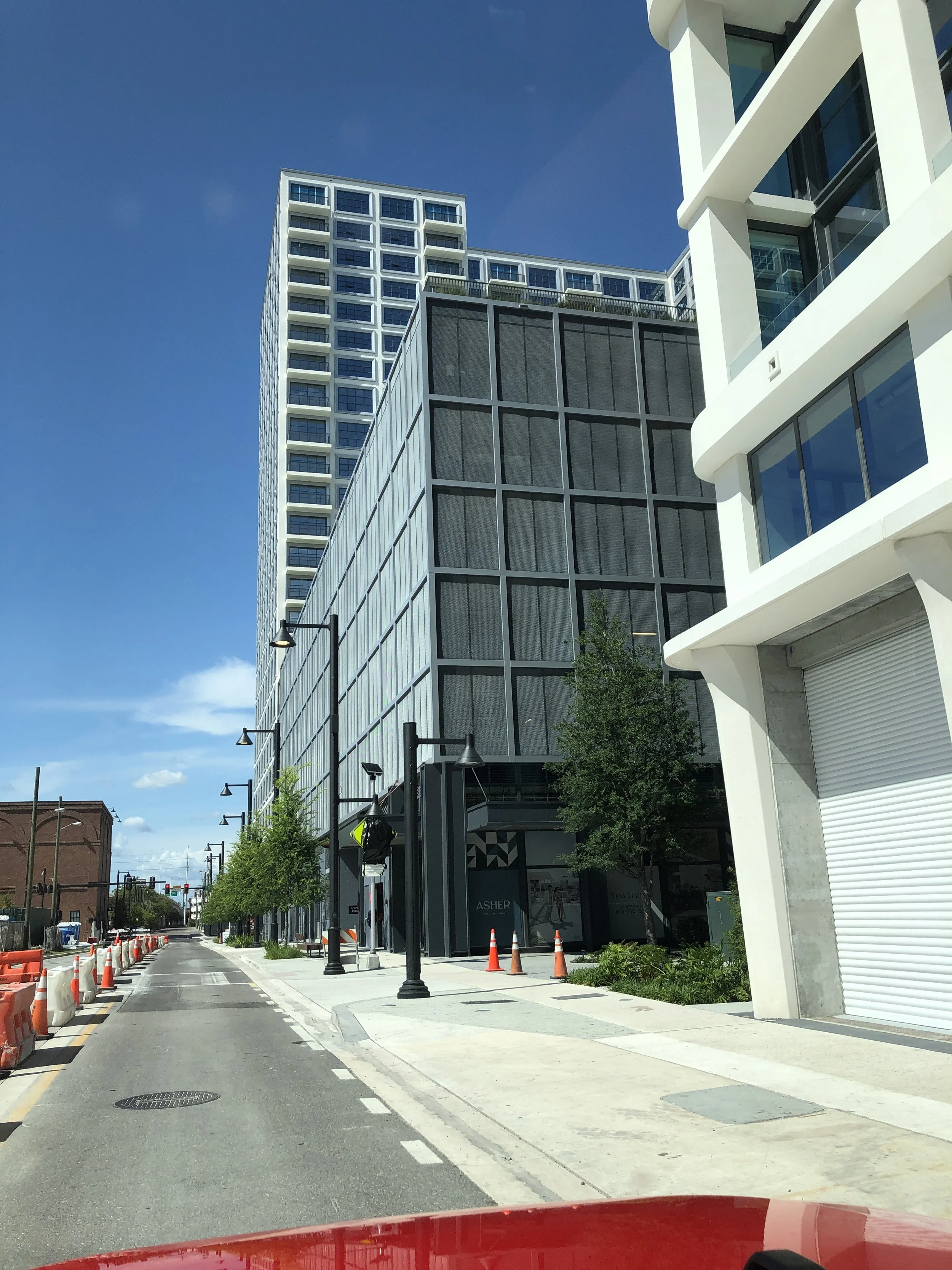 Urban street scene with modern high-rise buildings, construction cones, street lamps, trees, and clear blue sky.