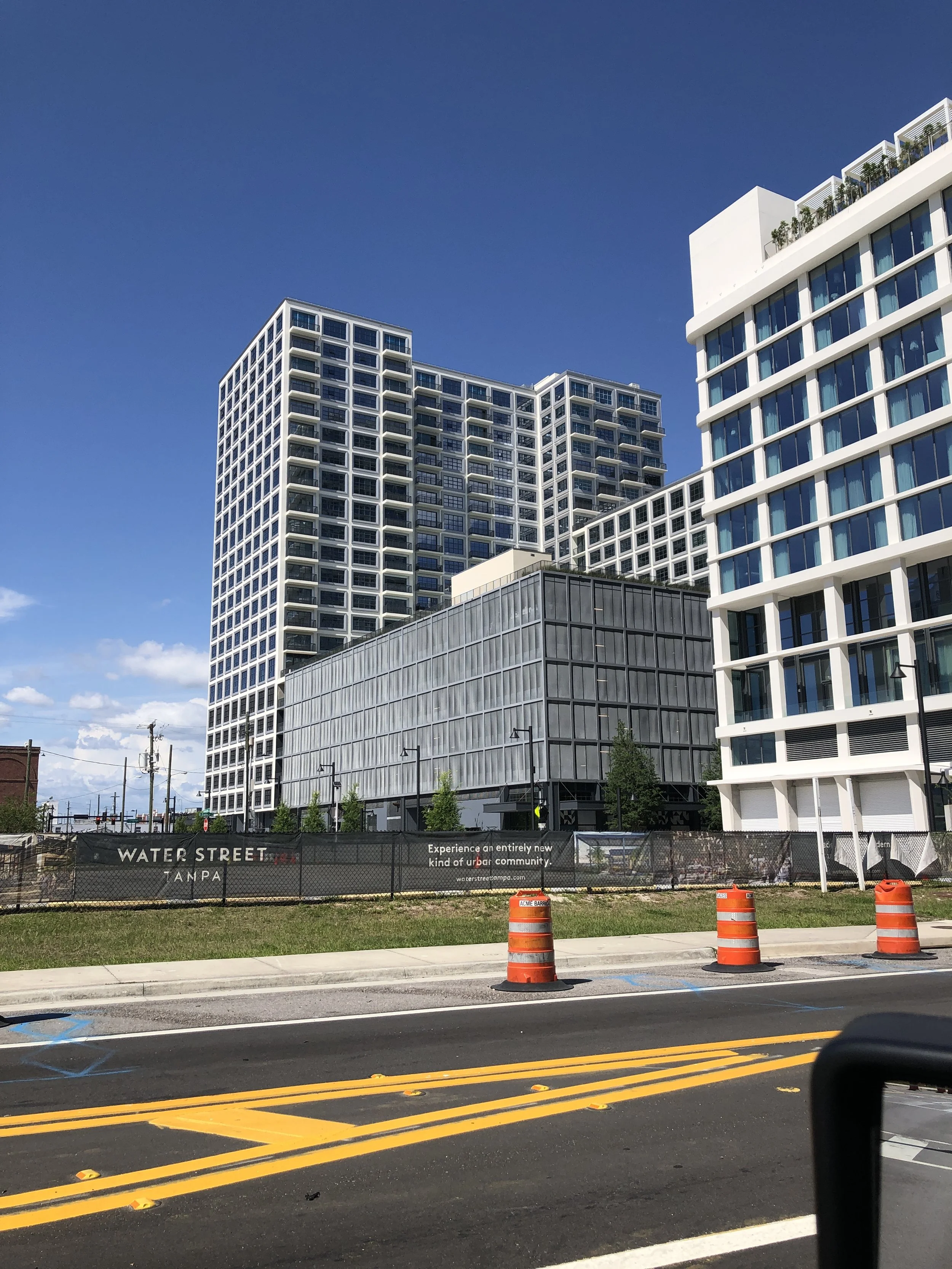 View of modern multi-story buildings under a clear blue sky, with a construction site and orange traffic barrels in the foreground.