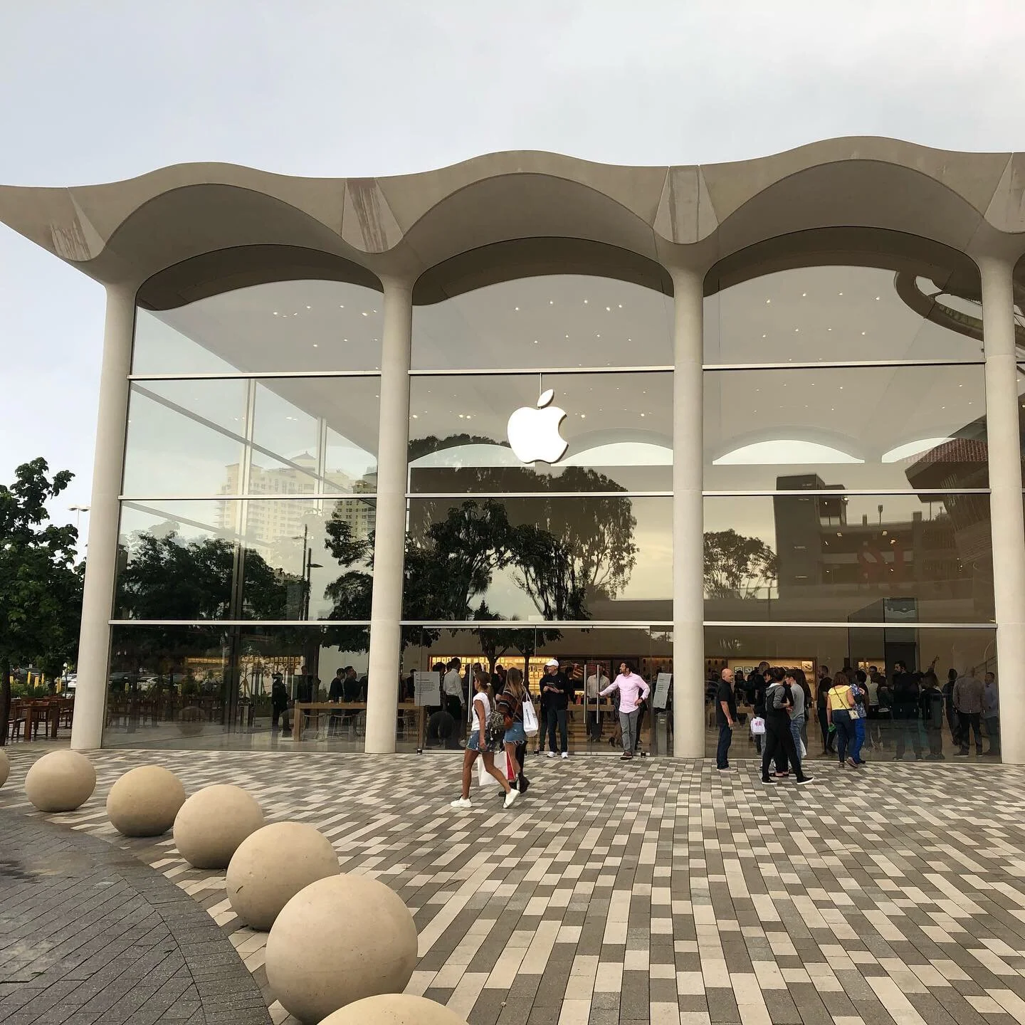 Exterior of an Apple Store with large glass windows, a white Apple logo, and people entering and exiting the store.