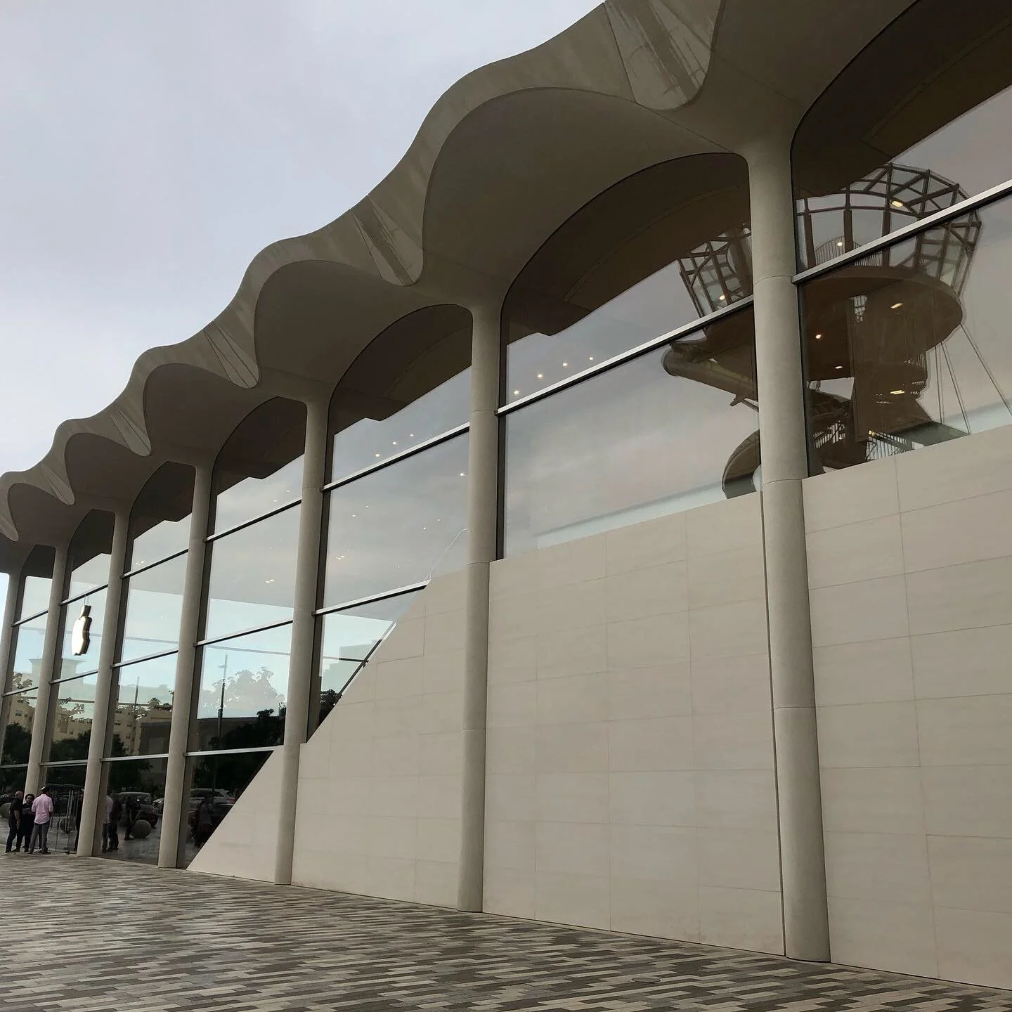 Modern building with curved roof and large glass windows, with a few people near the entrance, and a parking lot visible in the background.