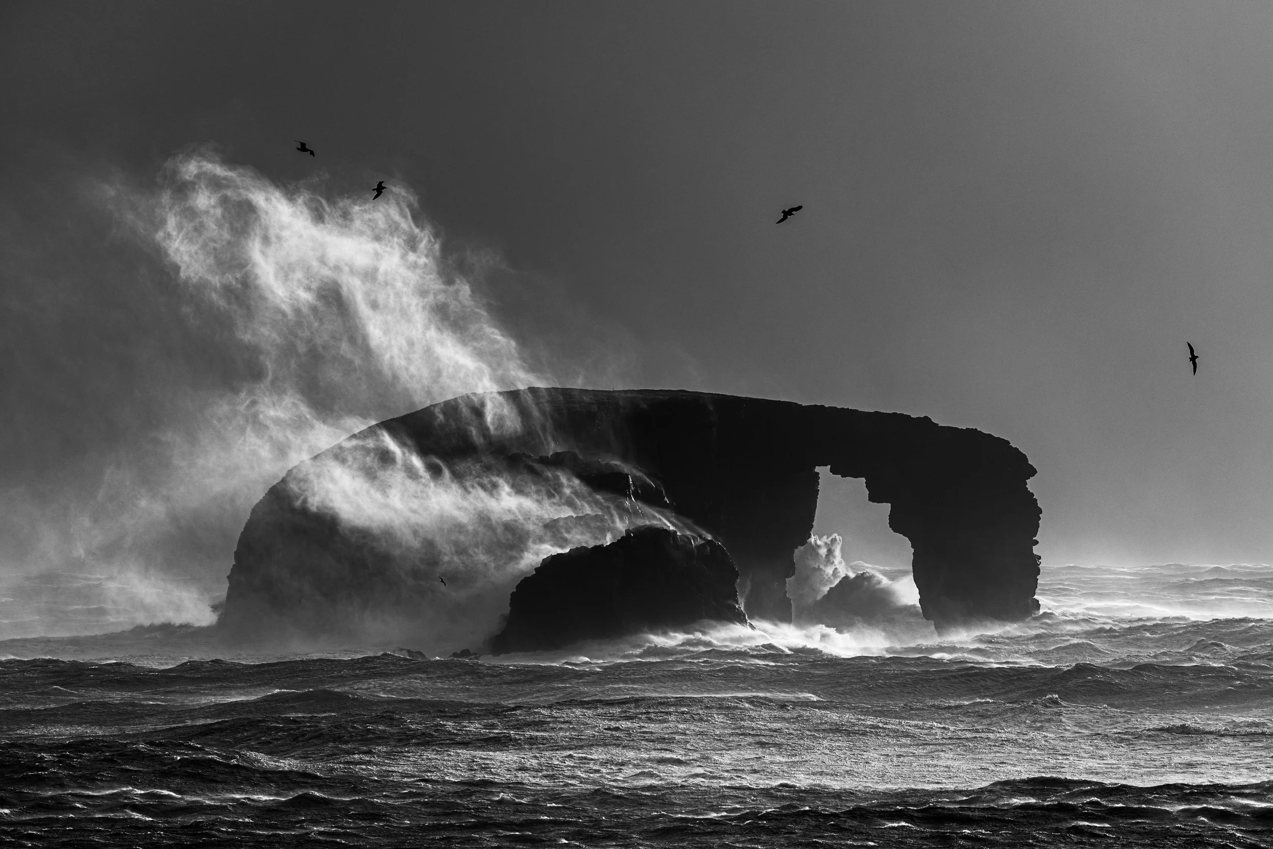 Black and white photo of a coastline with large rock formations and waves crashing against them.