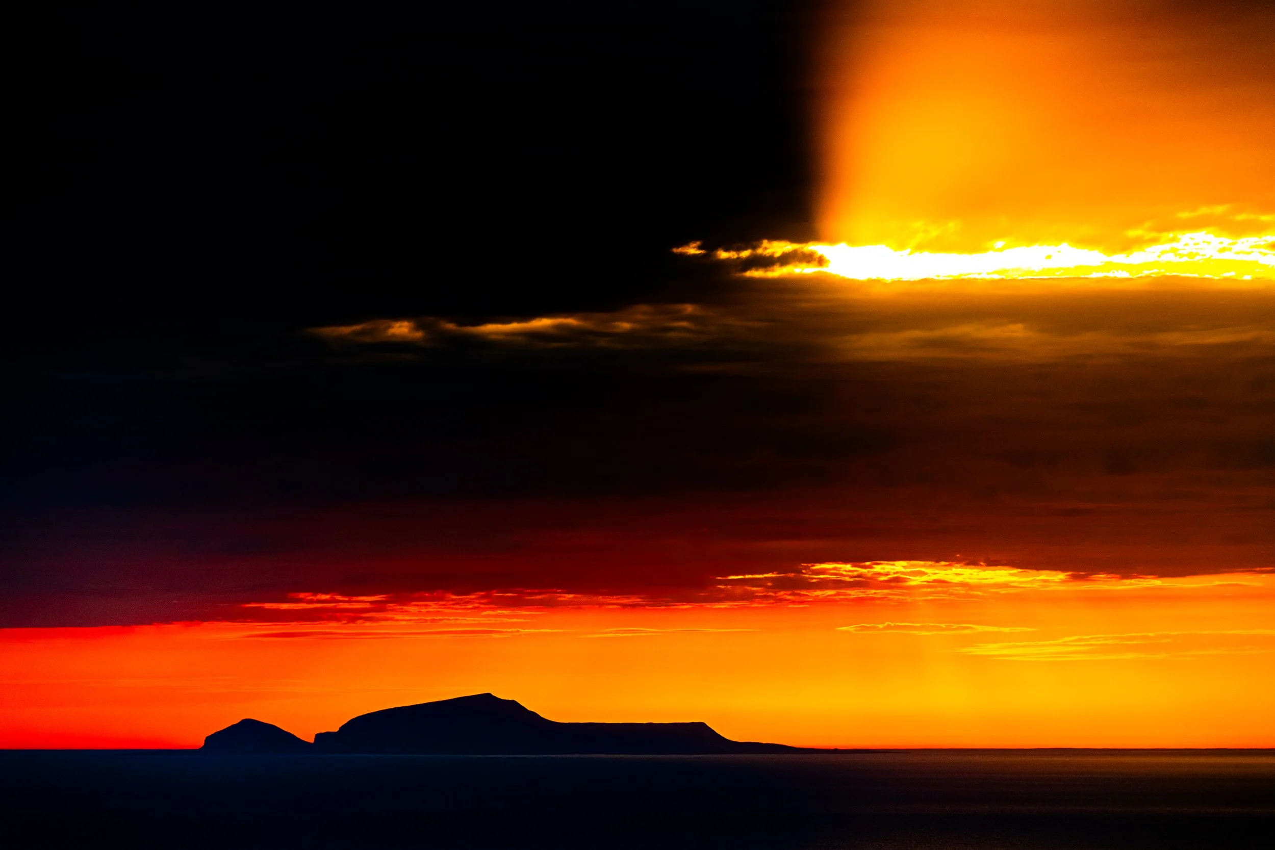 Sunset over the ocean with a silhouette of land and mountains, vibrant orange and yellow sky, dark clouds, and reflections on the water.