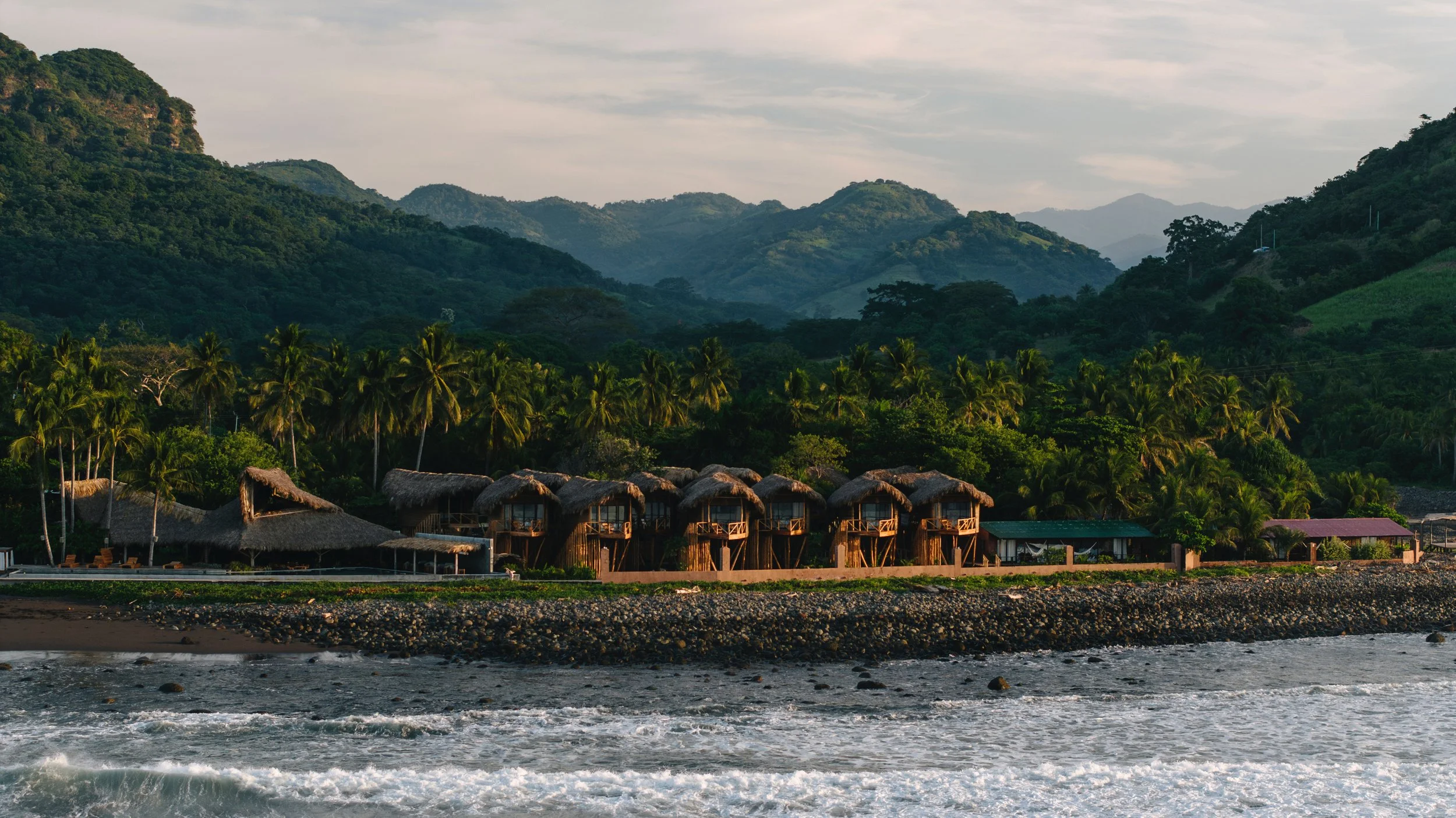 Beachfront view with a row of thatched-roof huts, lush green palm trees, and densely forested hills in the background under a partly cloudy sky.